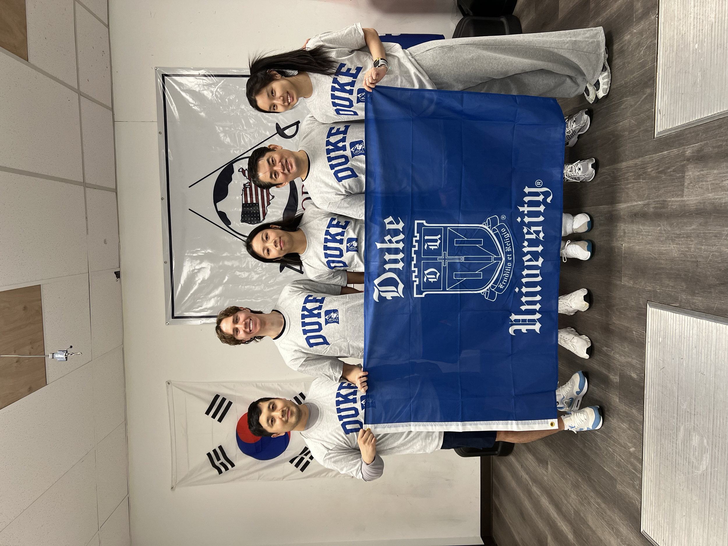 Four young adults standing together indoors, holding a blue flag with Duke University Athletic logo, with the South Korean flag hanging on the wall behind them.
