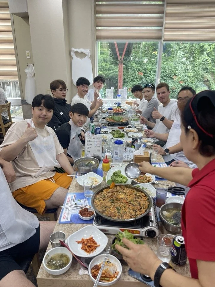 A group of people sitting around a long table enjoying a meal at a restaurant, with various dishes and drinks, during daytime.