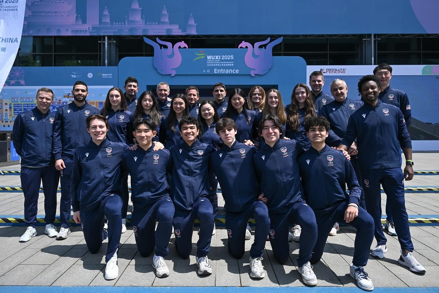 A large group of athletes in matching navy blue tracksuits with team logos pose for a photo outside a stadium entrance with a sign that reads 'WUXI 2025' and features stylized dragons and a castle silhouette. The group includes men, women, and childr