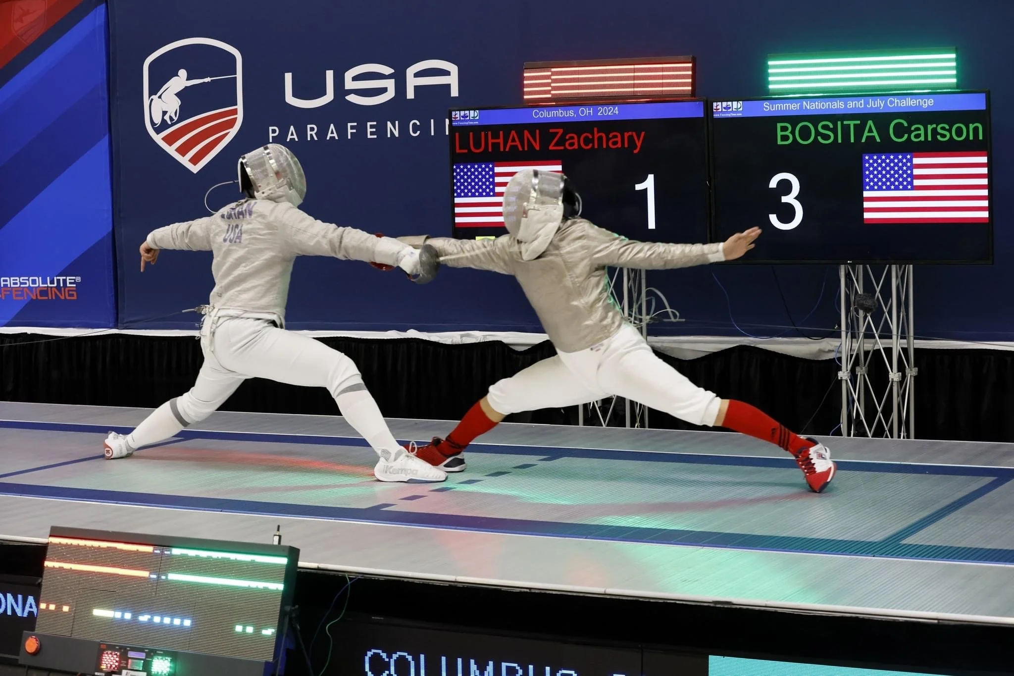 Two fencers from the USA competing in a fencing match, engaging in a lunge with their swords extended towards each other. They are on a fenced strip with electronic scoring equipment. A scoreboard shows the names 'LUHAN Zachary' and 'BOSITA Carson,' 