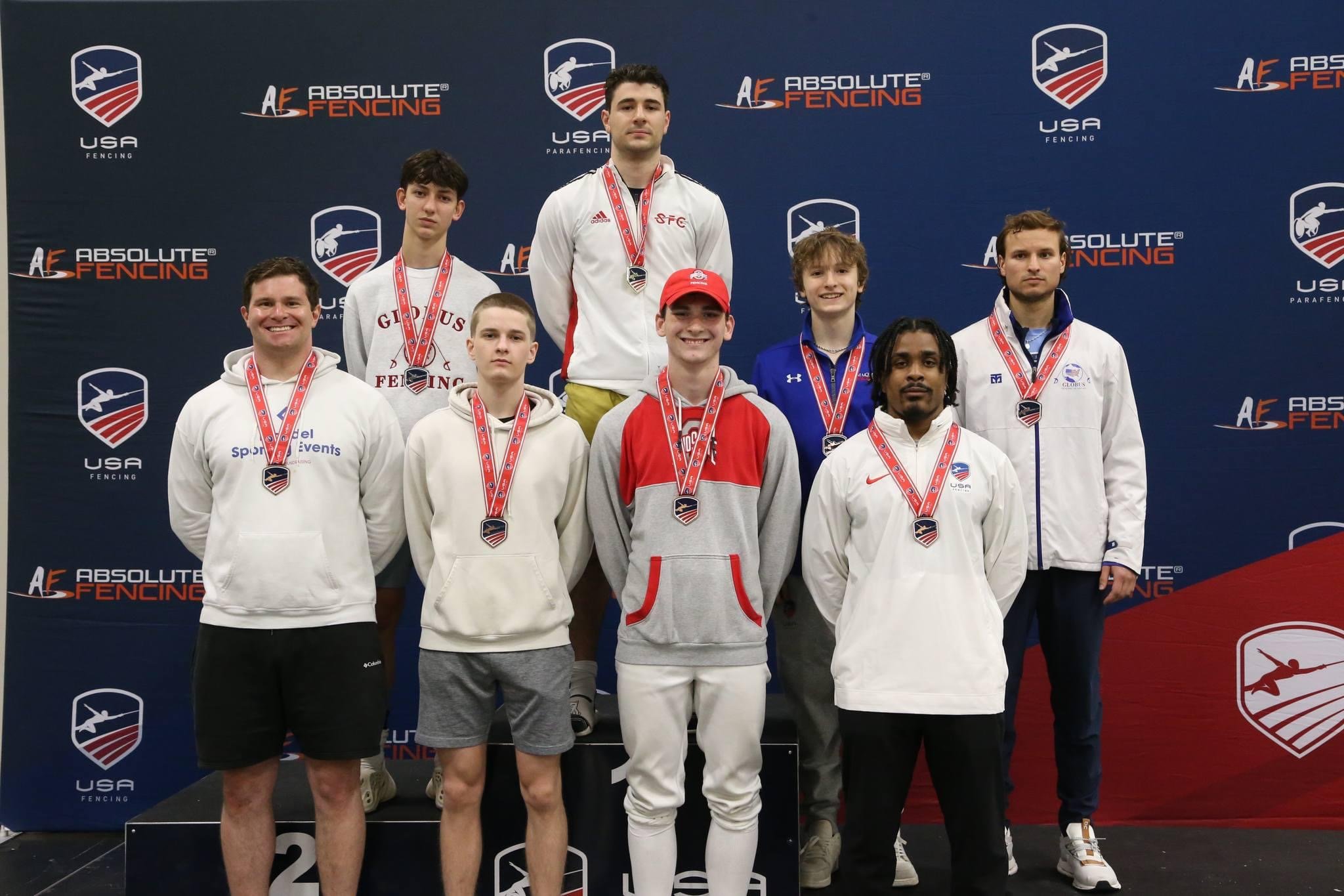 Group of ten young male fencers standing on a podium and stage, wearing medals and sportswear, at a fencing competition with banners for USA Fencing and Absolute Fencing in the background.
