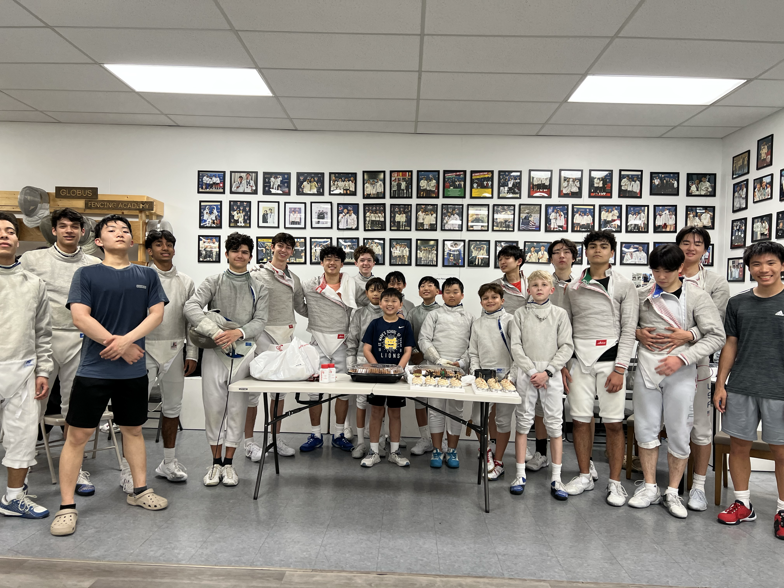 Group of young people dressed in fencing attire standing around a table with food and drinks, in a room with walls decorated with sports photos.