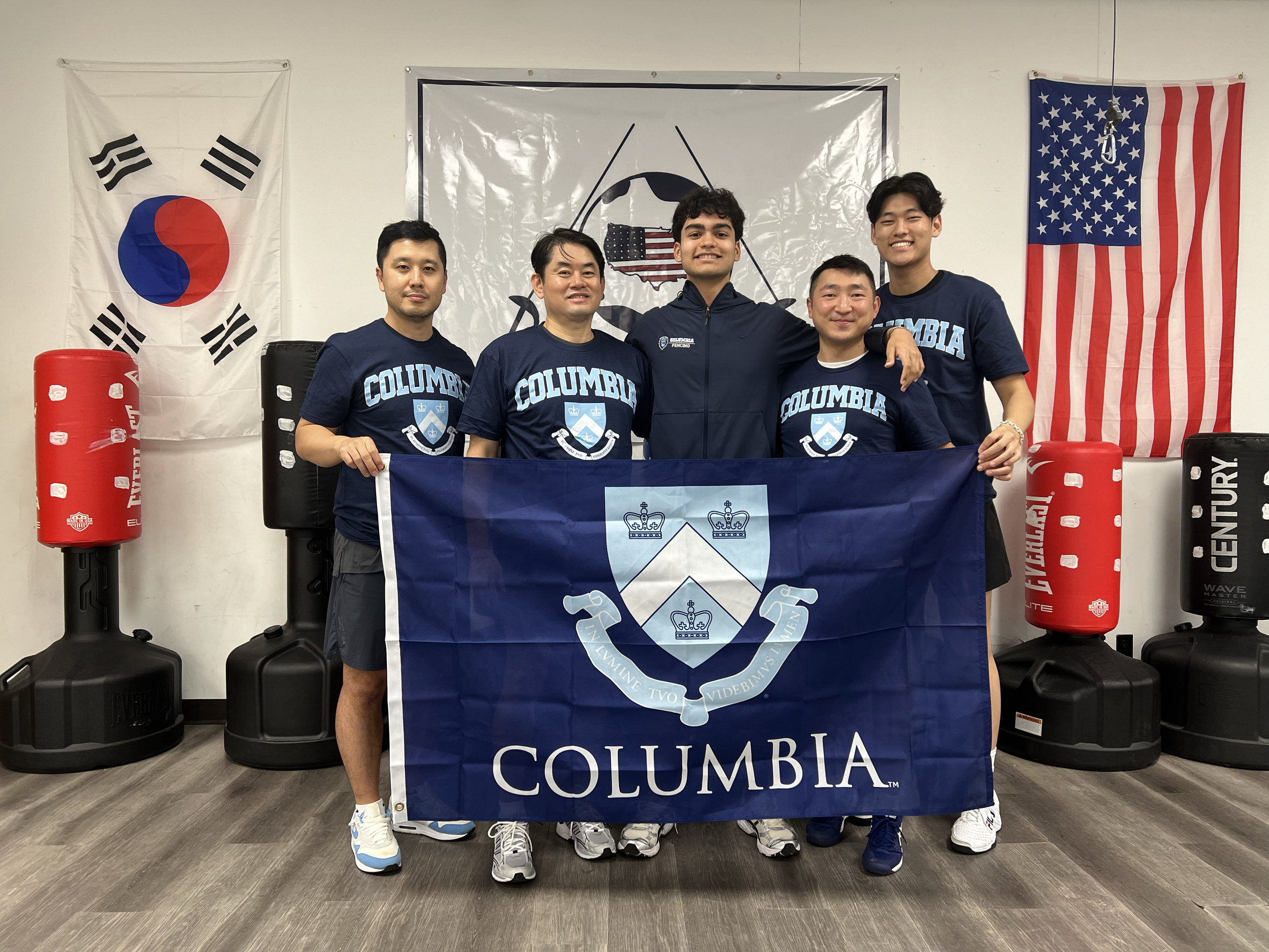 Group of five young men in Columbia University shirts holding a Columbia banner, standing in a martial arts gym with flags of South Korea and the United States in the background.