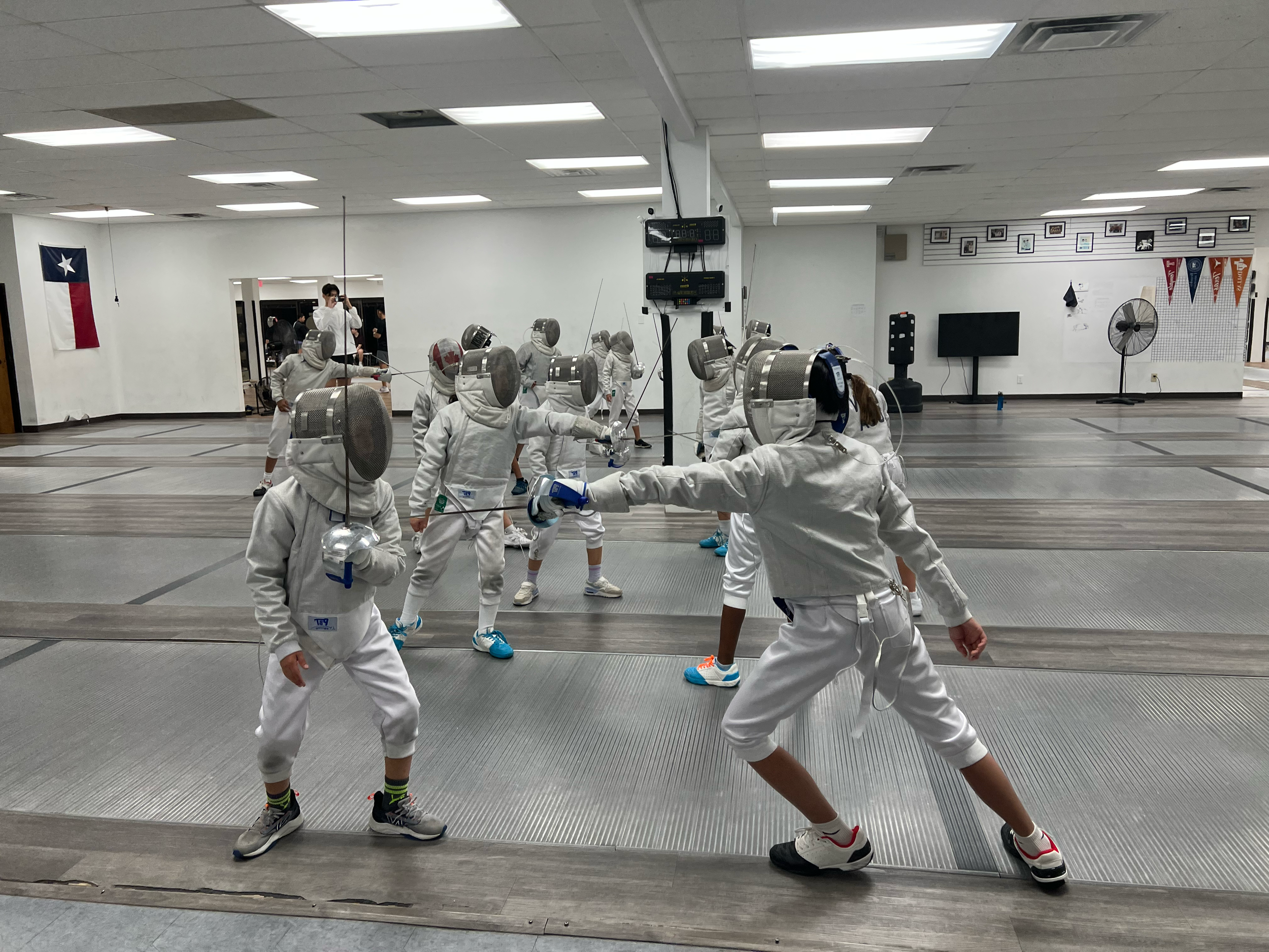 Children and instructor practicing fencing indoors, all in full fencing gear including masks, jackets, and gloves, on a fencing strip.