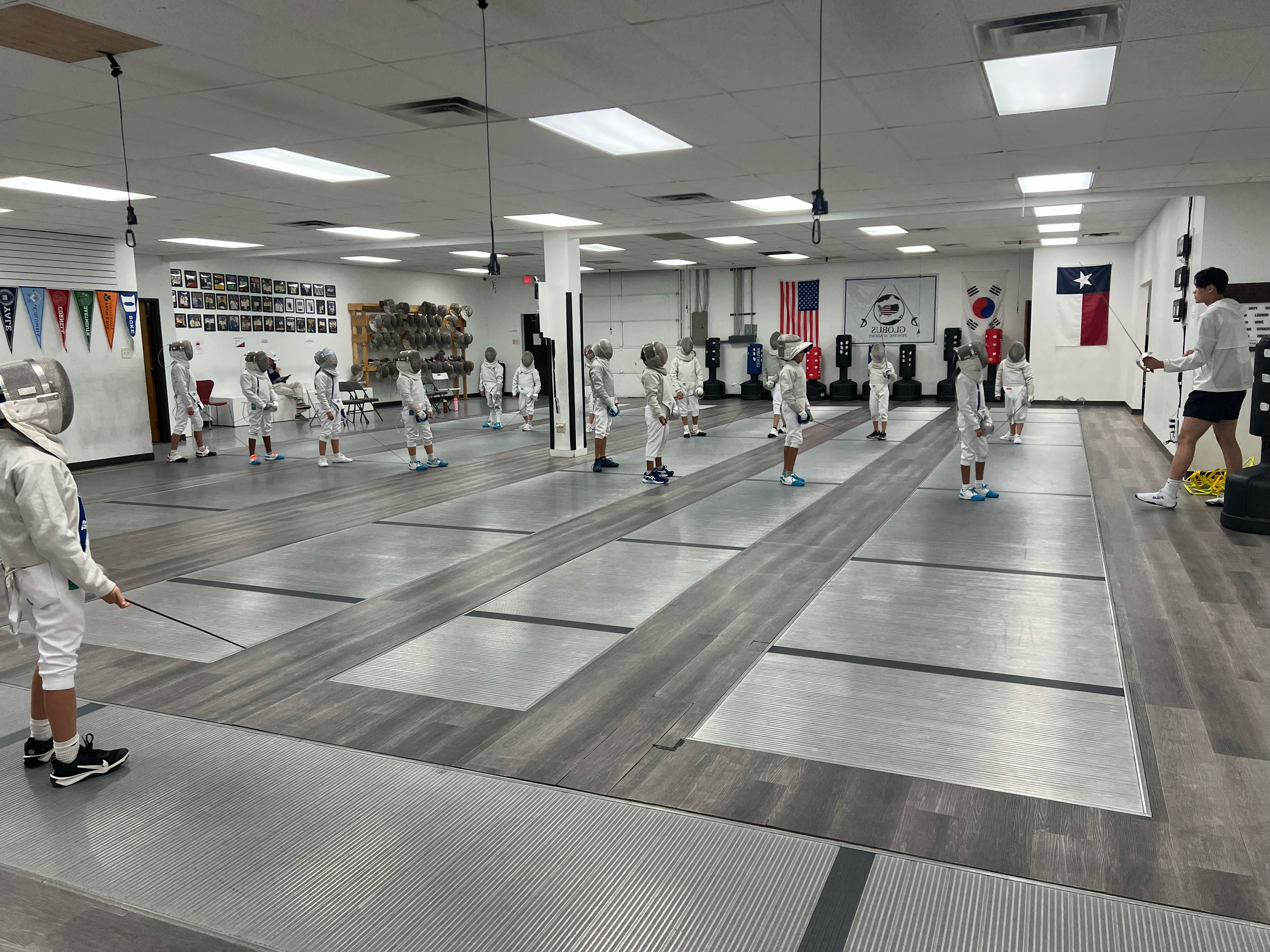 Children practicing fencing under supervision of instructor in a fencing studio with flags and wall decorations.