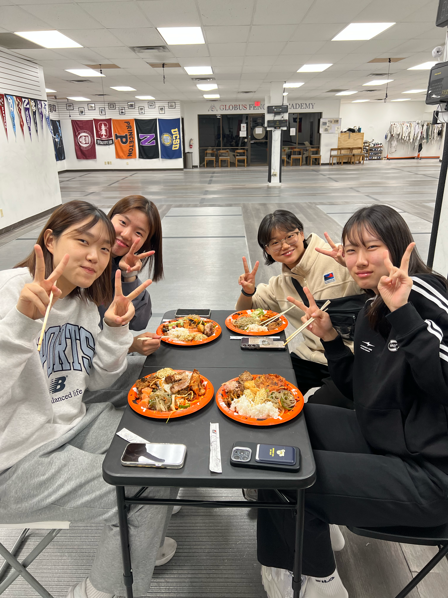Four young women sitting at a table with orange trays of Asian cuisine, making peace signs and smiling inside a martial arts gym. Walls are decorated with flags and banners.