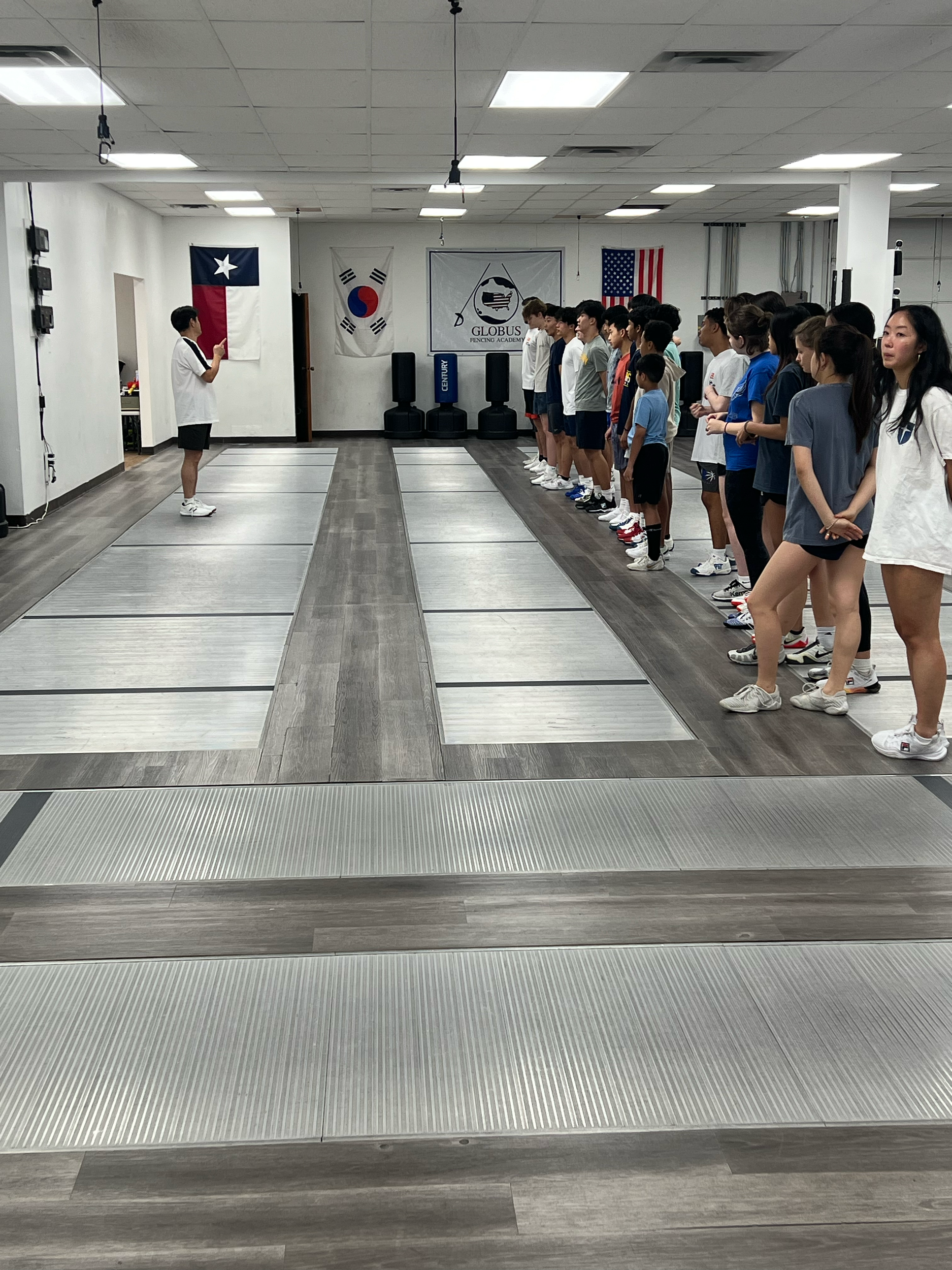 Instructor leading a fencing class with students lined up in a martial arts training room decorated with flags including the Texas, South Korea, and United States flags.