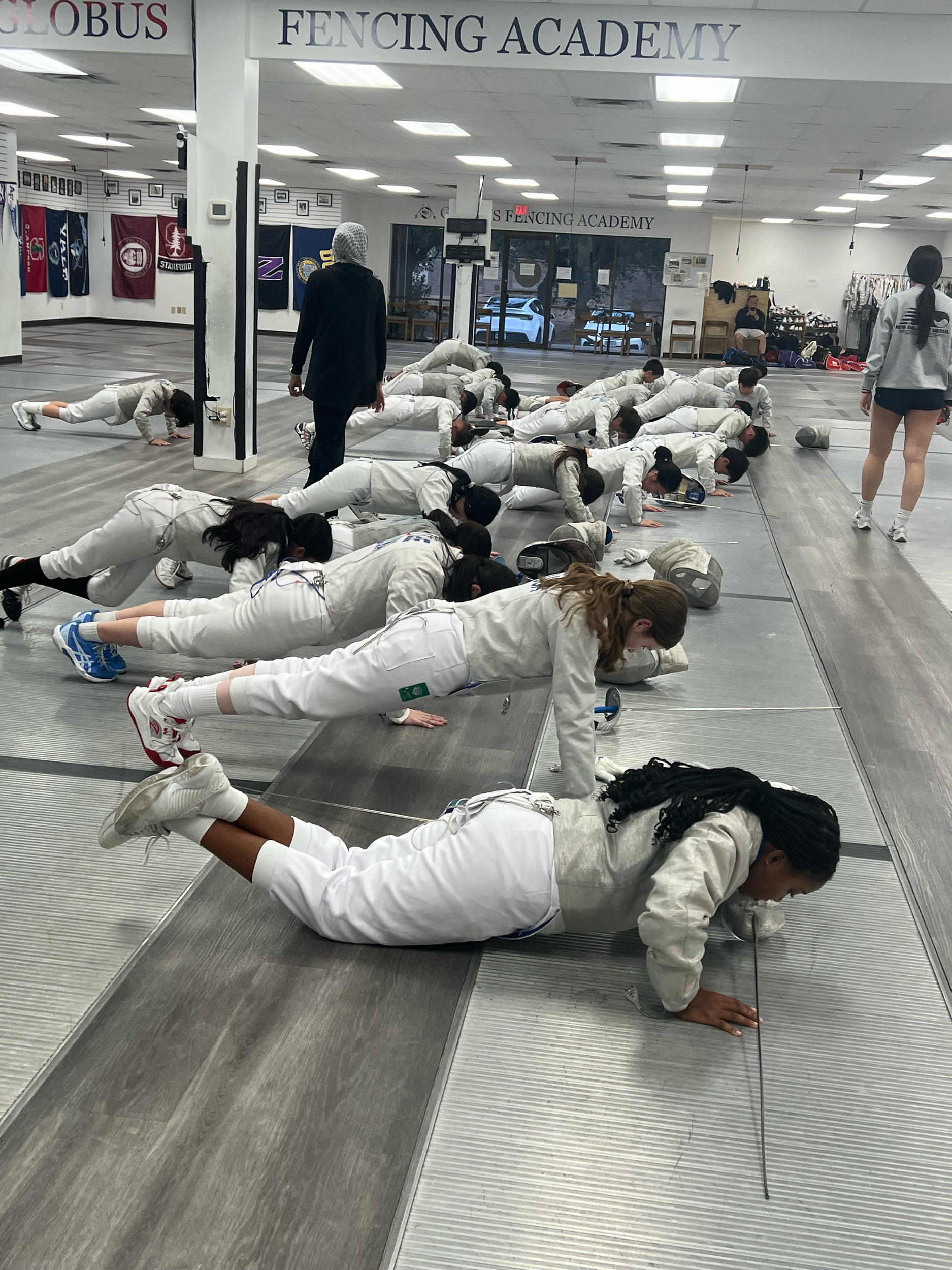 A group of students in fencing uniforms practicing push-ups on the ground inside a fencing academy. The room has banners, flags, and high ceilings.