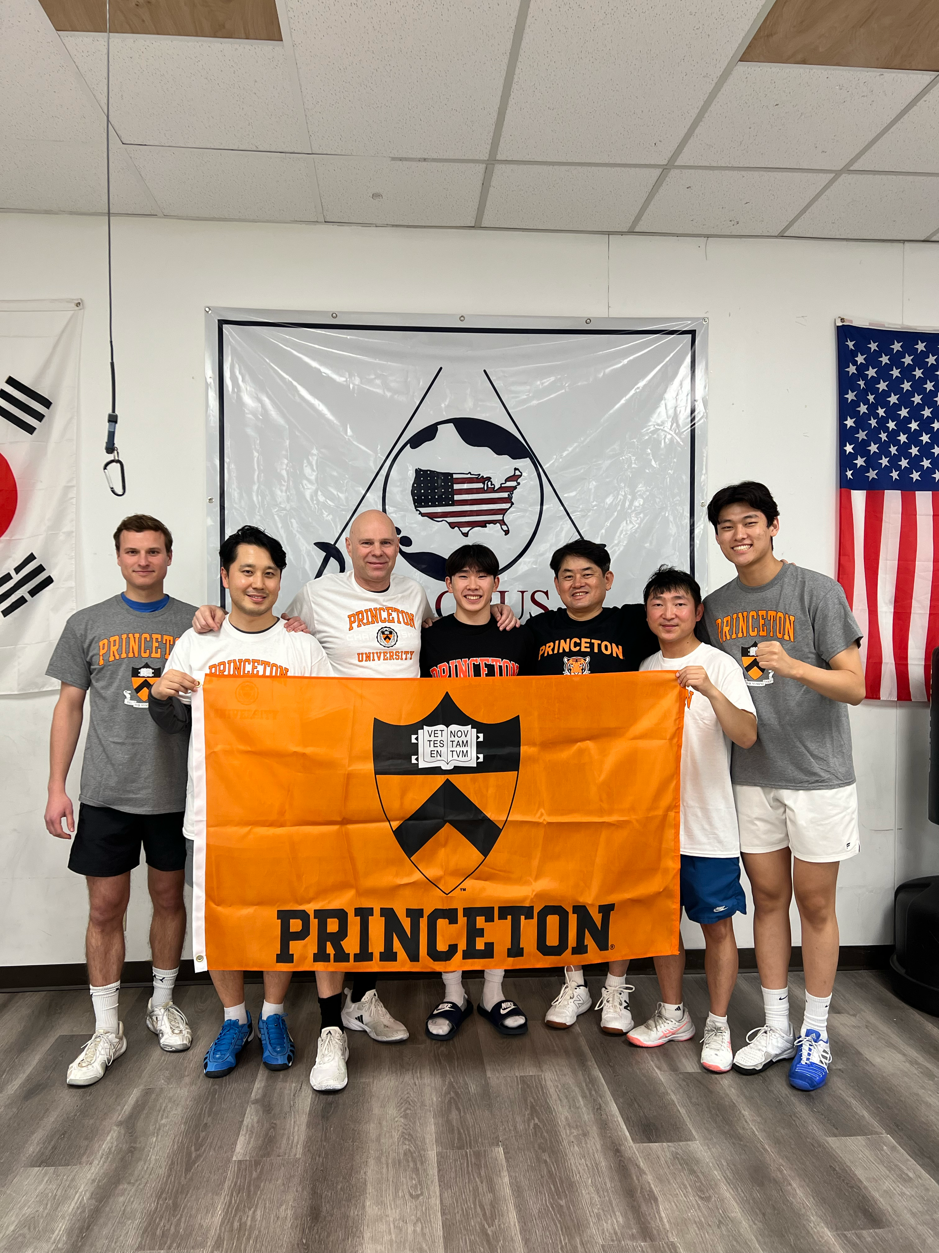 Group of seven people in Princeton University apparel holding an orange Princeton flag, standing in front of flags including South Korea and United States, and a banner with an eagle and shield emblem on the wall.