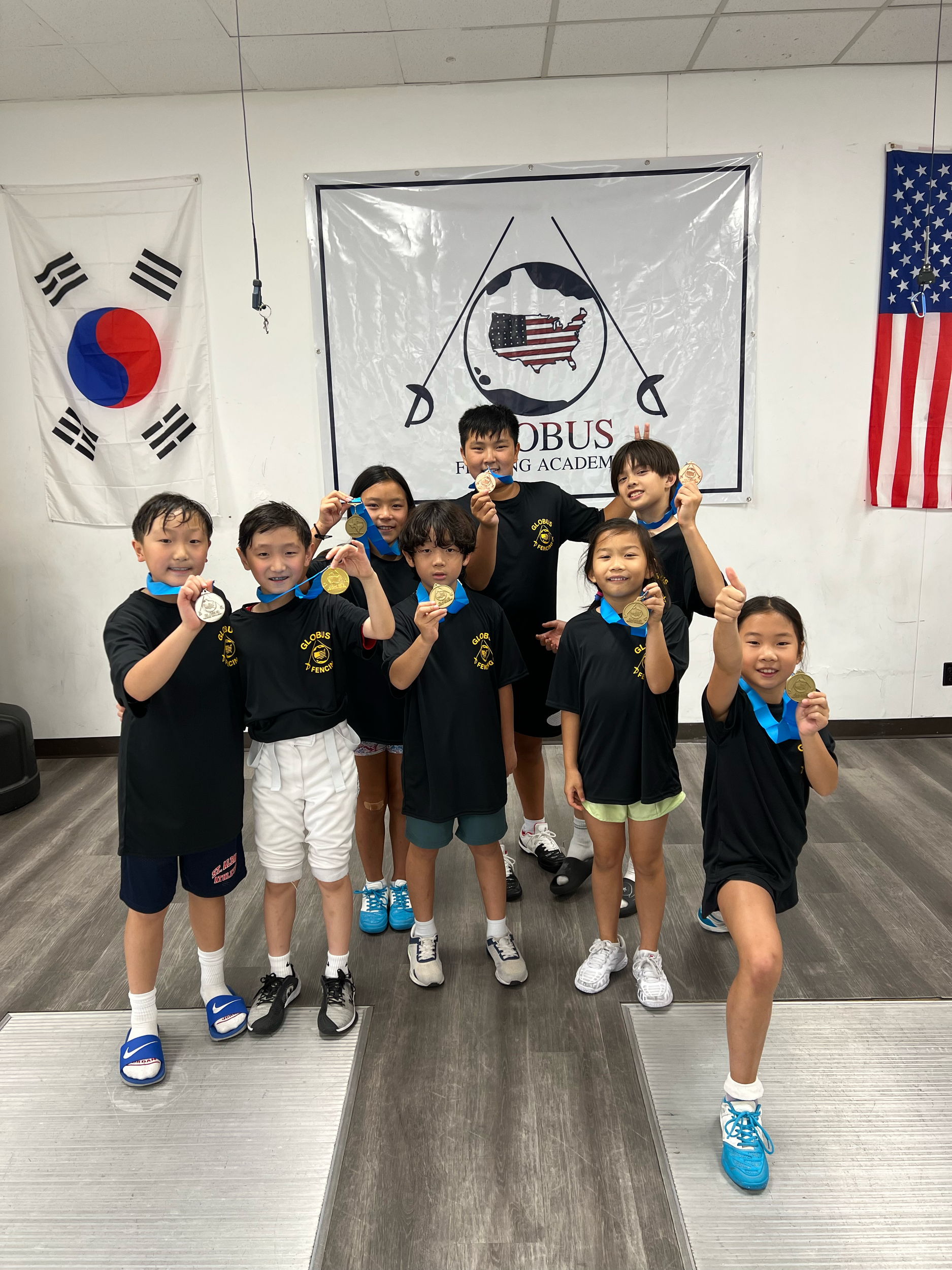 Children standing in a row on a podium holding medals after a fencing competition, with South Korean and American flags hanging on the wall behind them.
