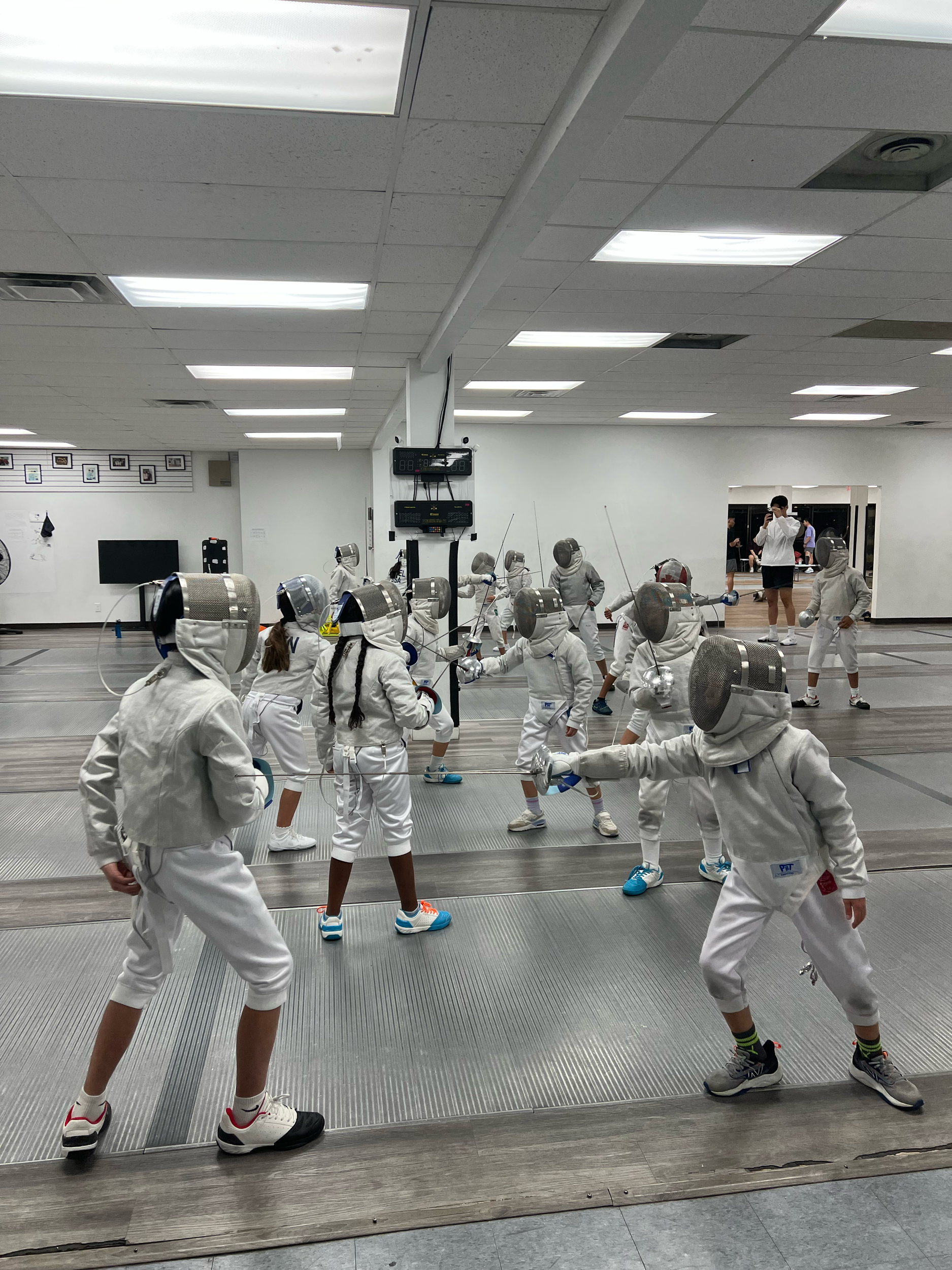 Children practicing fencing in an indoor fencing facility, wearing protective gear and masks.