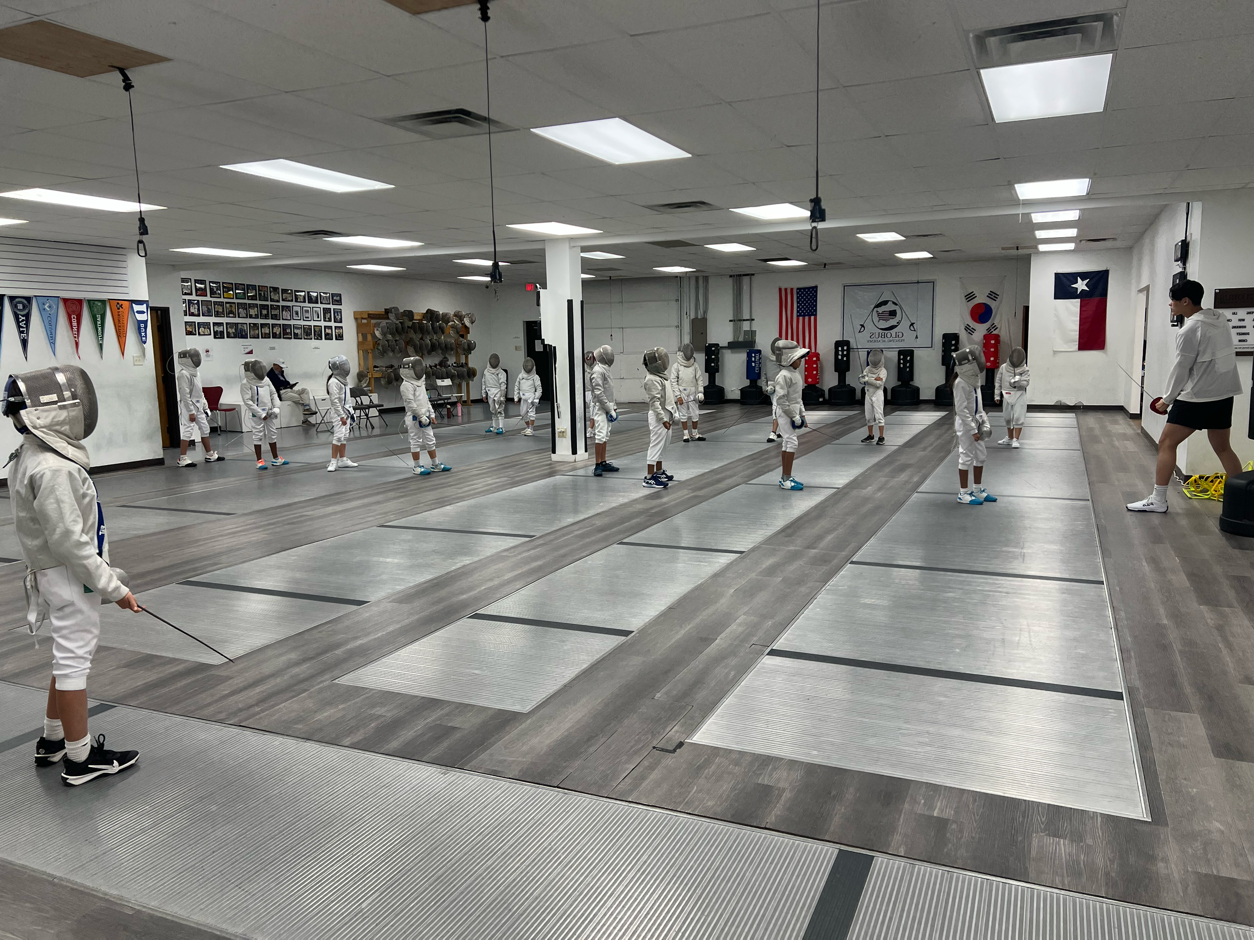 Children in fencing protective gear practicing fencing indoors, being instructed by a coach, with flags and photos on the walls.