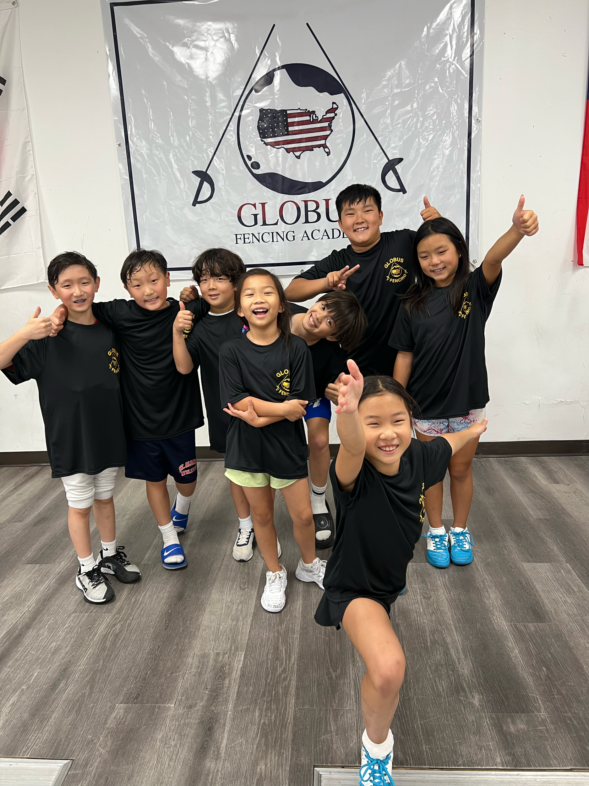 Group of children wearing black Globus fencing academy t-shirts, smiling and posing cheerfully in front of a banner with the academy's logo and a map of the United States.