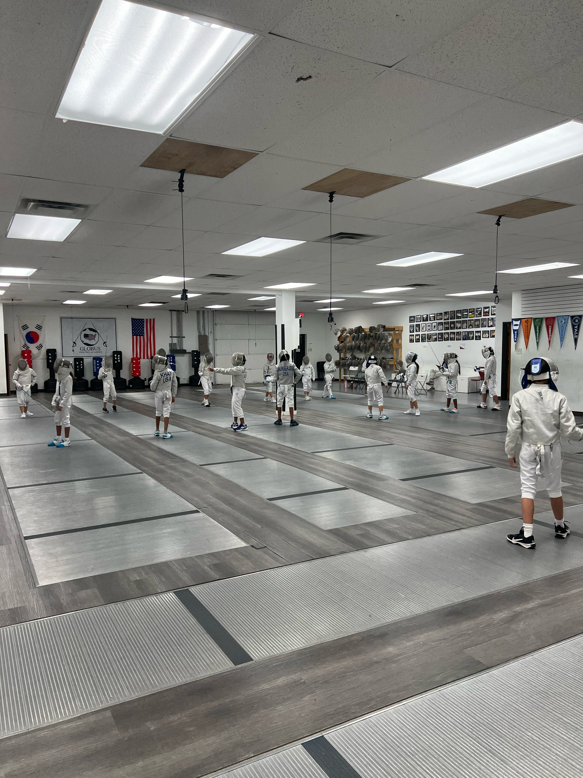 Youth fenced fencing class in a fencing gym, with students in white fencing gear practicing. The room has lined fencing lanes, flags, and martial arts memorabilia on the walls.