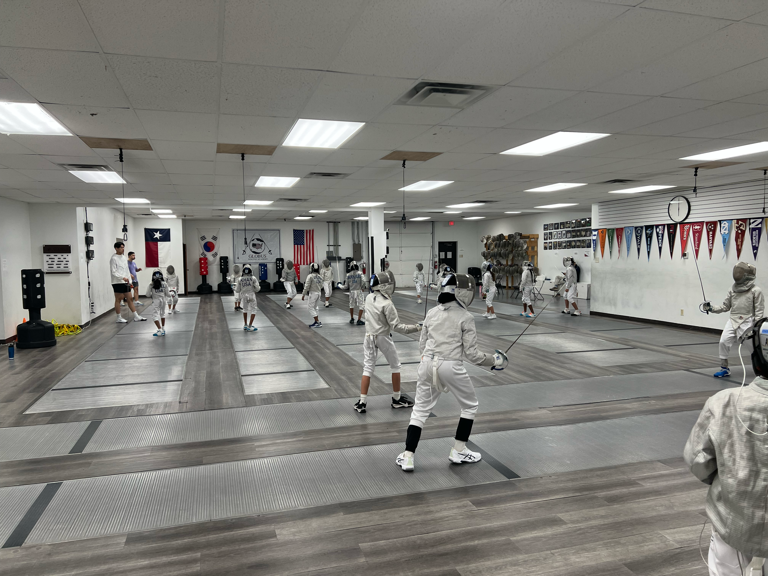 People practicing fencing in an indoor fencing gym with flags and photos on the walls.