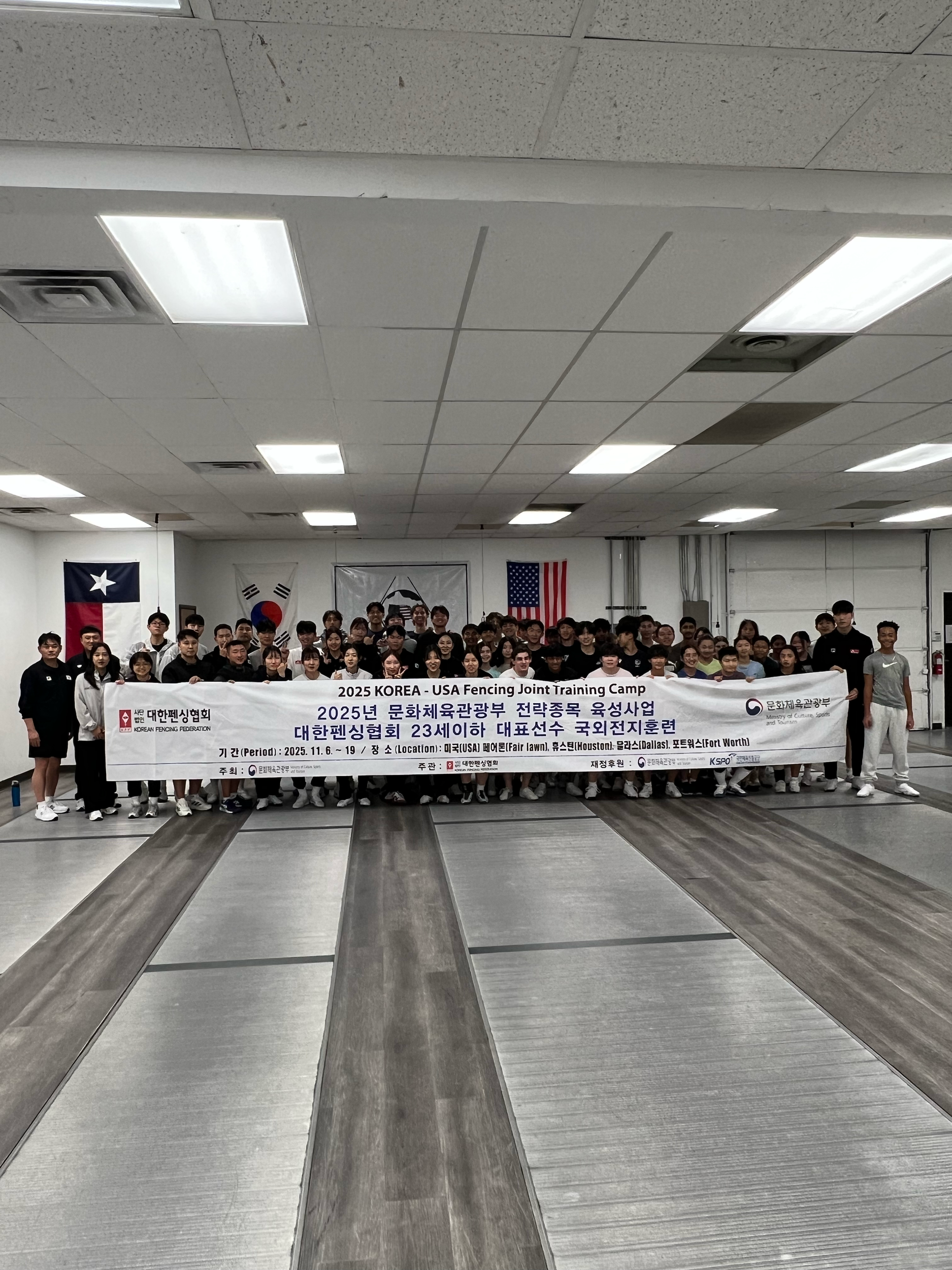 Group of people holding a large banner inside a fencing gym, with flags of Texas, South Korea, and the United States hanging on the wall behind them.