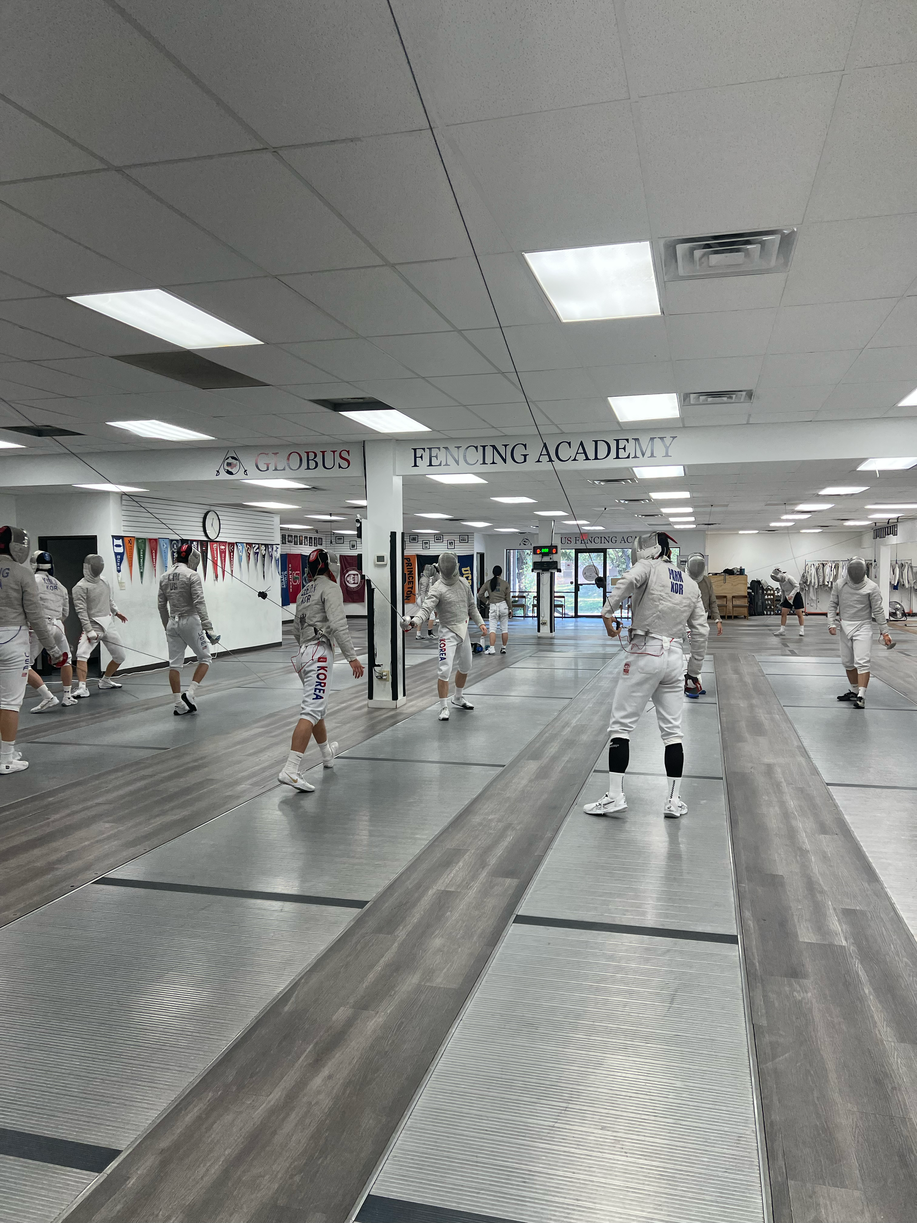 Indoor fencing training facility with people practicing fencing, mannequins wearing fencing gear, and banners on the wall.
