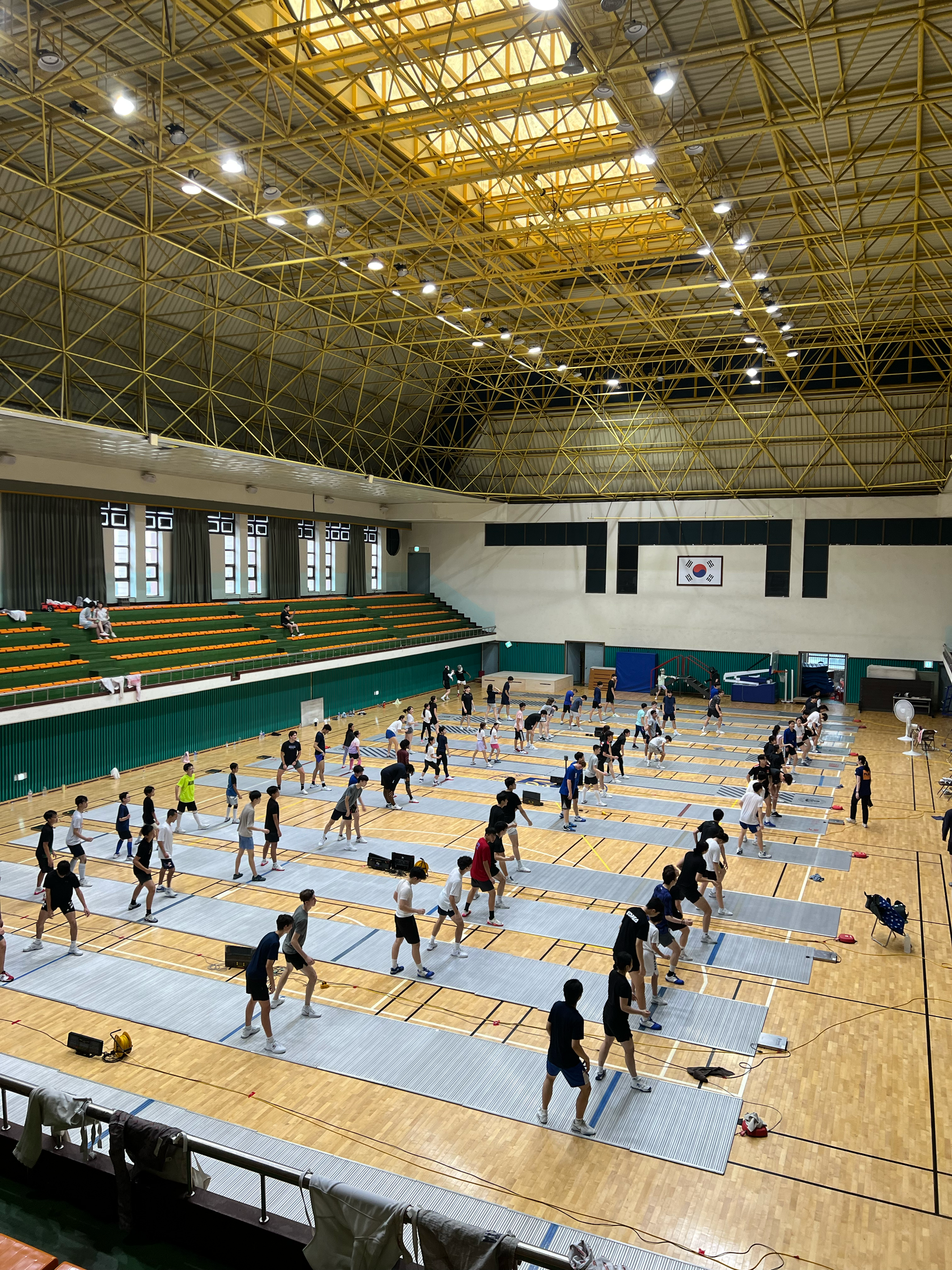 People participating in a group workout or exercise class in an indoor gymnasium with a Korean flag on the wall.