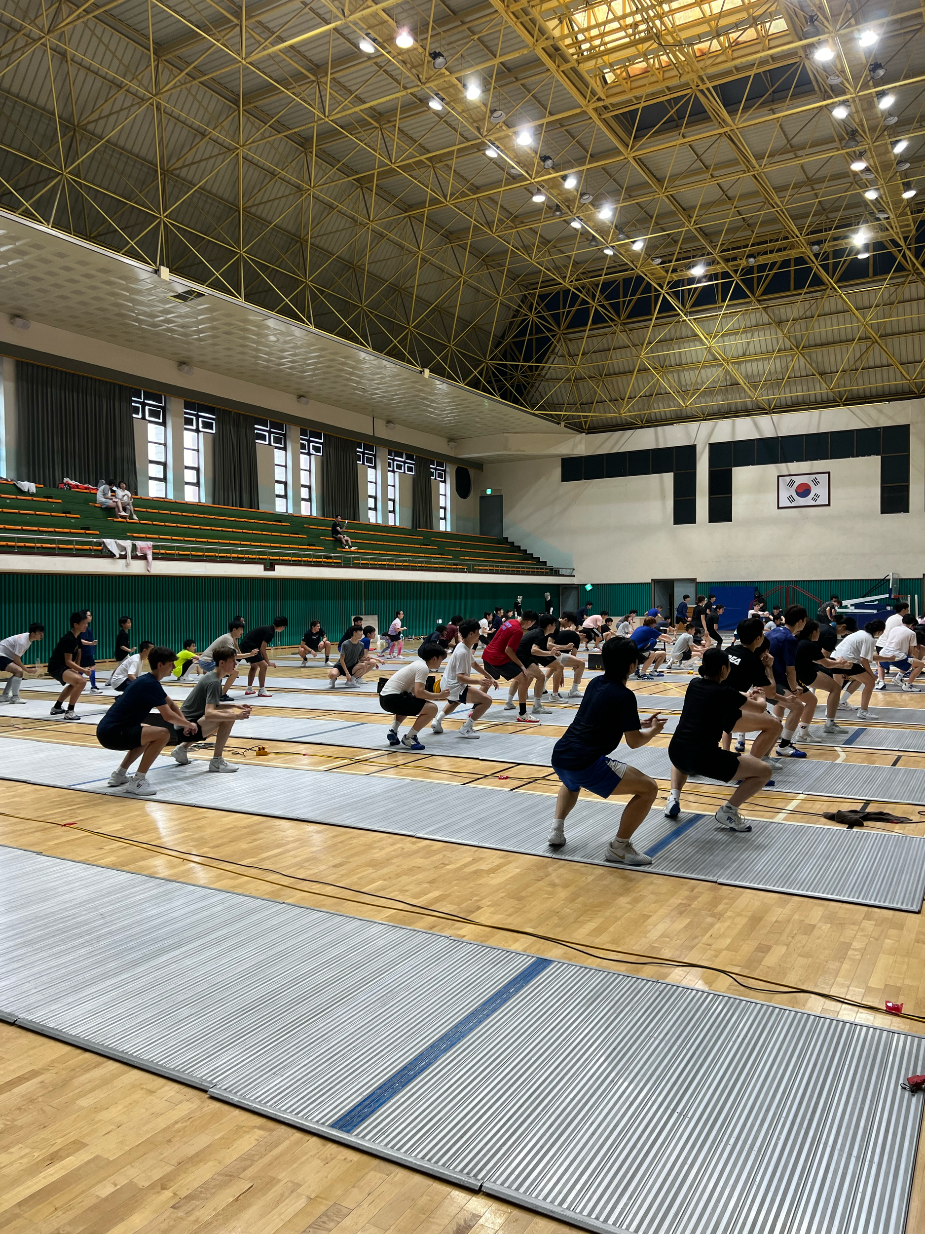 Group of people participating in a fitness class or exercise session in a gymnasium with high ceiling, large windows, and the South Korean flag on the wall.