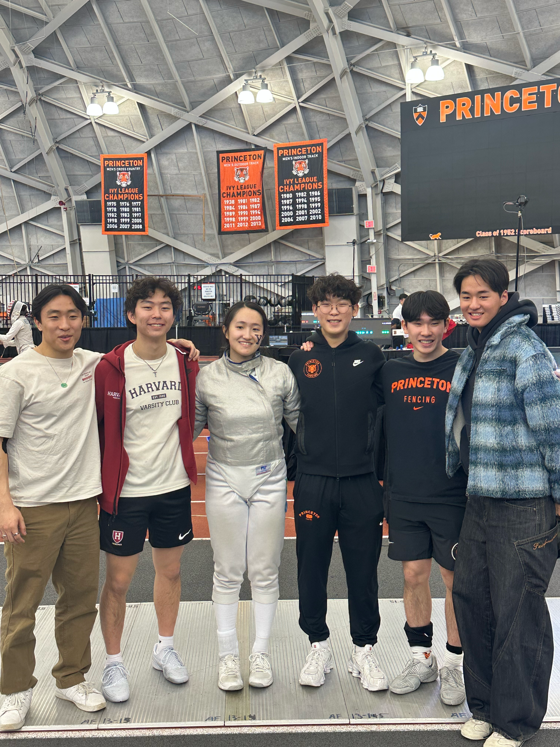 Group of six young people posing inside an indoor athletic stadium, with banners and a scoreboard in the background, celebrating their fencing achievements.