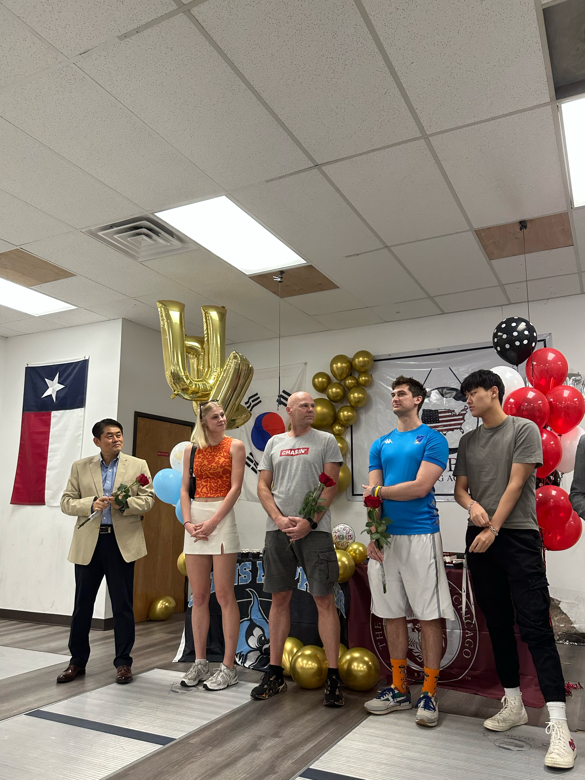 Group of five people at a celebration with balloons, a large gold balloon spelling 'U', a Texas flag on the wall, and a decorated backdrop with red, black, gold, and white balloons.
