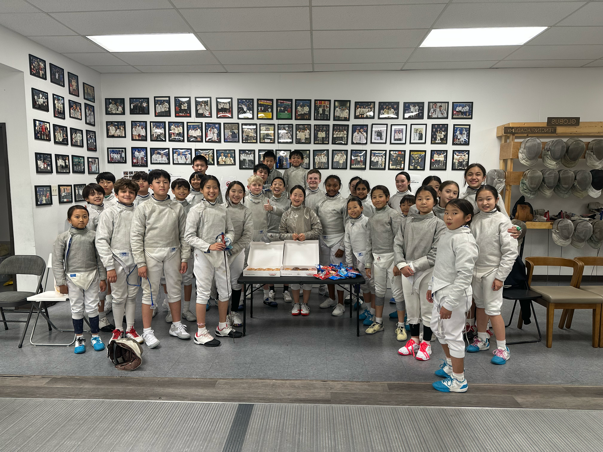 A group of children in fencing uniforms posed indoors, standing around a table with snacks and pizza, with framed photos on the wall behind them.