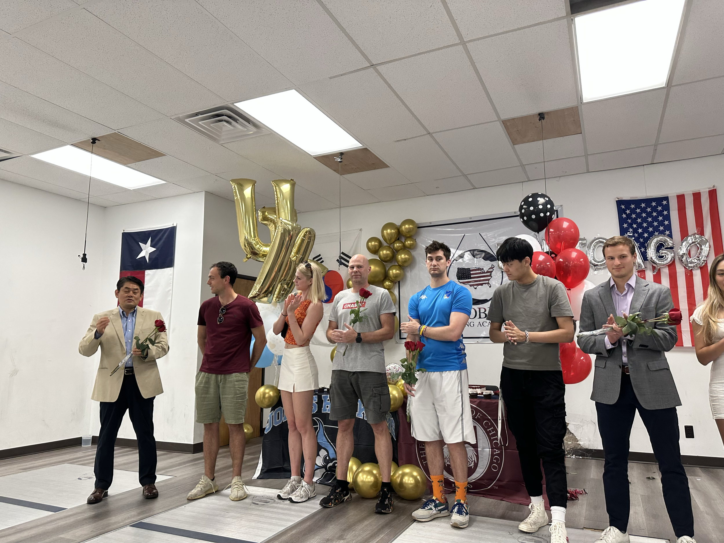 Group of seven young adults standing on a stage at an indoor event, holding roses and engaging in a ceremony or celebration, decorated with balloons, American flags, and banners, with a Texas flag on the wall.