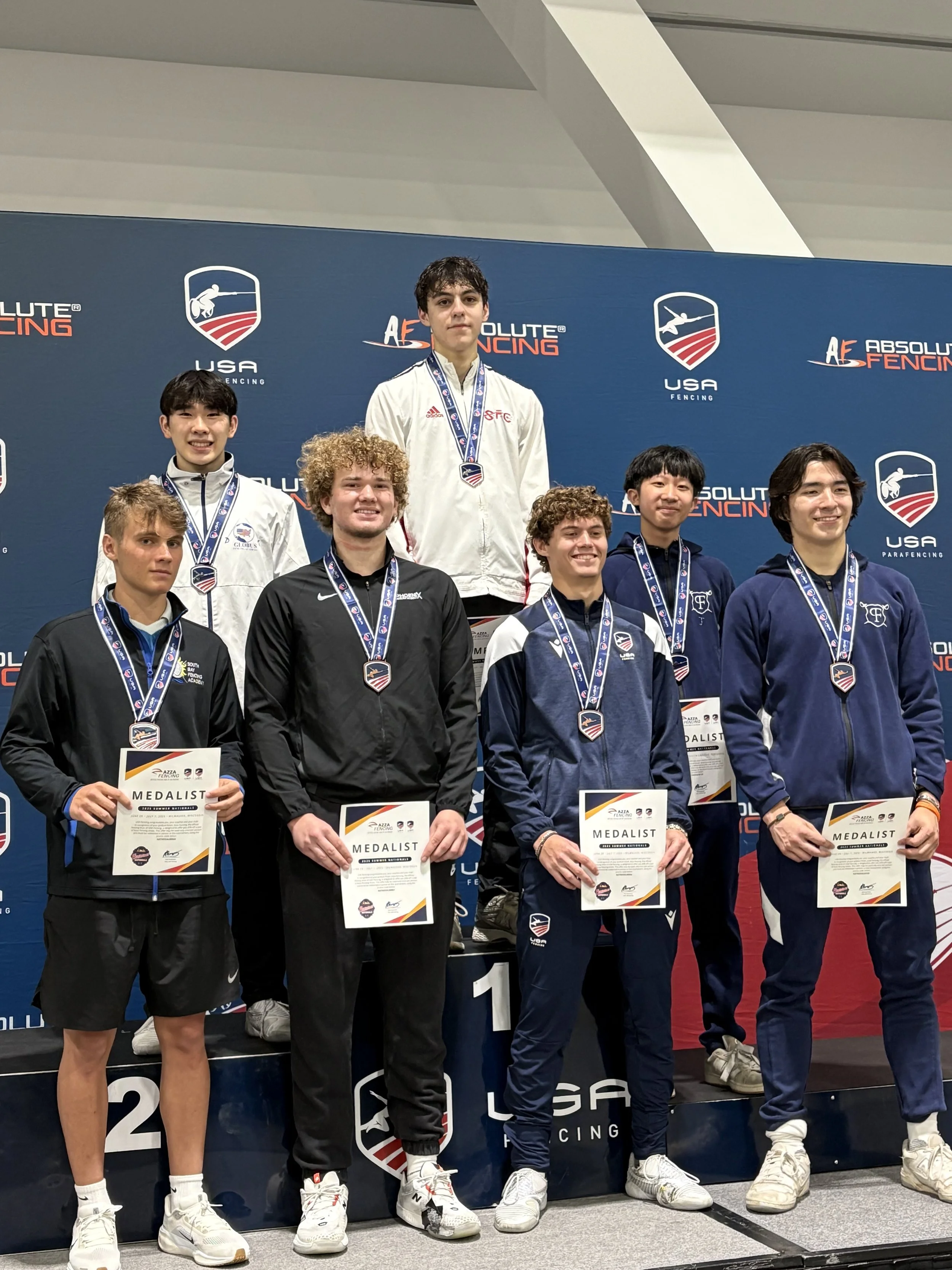 Young fencers standing on a podium after a competition, holding medals and certificates, with a backdrop displaying 'Absolute Fencing' and 'USA Fencing' logos.
