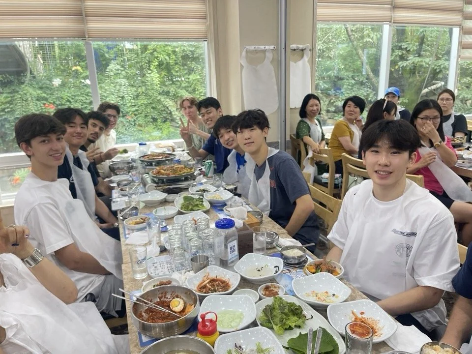 Group of young people and adults seated at a long dining table filled with plates of food, bowls, and drinks, celebrating together in a room with large windows showing greenery outside.