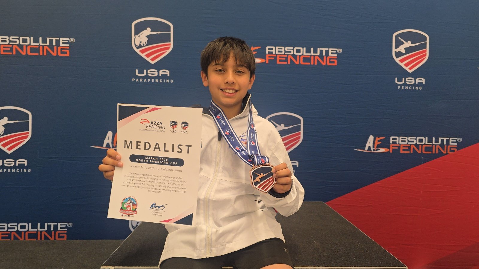 Young boy smiling and holding a medal and certificate at a fencing competition, with a backdrop displaying logos for Absolute Fencing and USA Fencing.