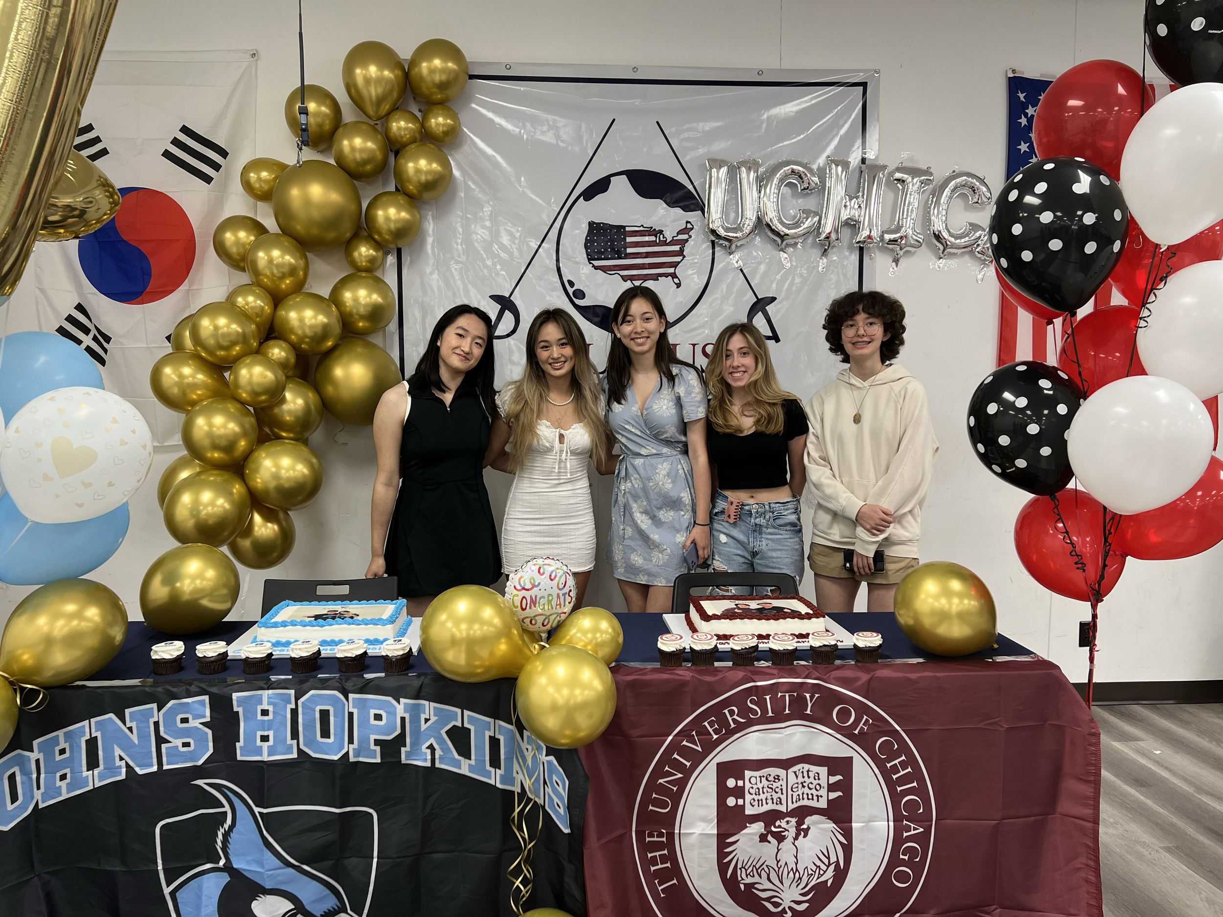 Group of five young women standing behind tables decorated for graduation or celebration, with banners and balloons from John Hopkins University and the University of Chicago, including a cake, cupcakes, and congratulatory decorations.