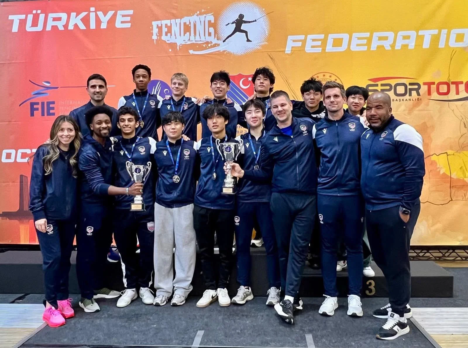 A group of athletes and coaches posing on a podium after a fencing competition, holding trophies and medals, with a large colorful banner reading 'Turkey Fencing Federation' in the background.