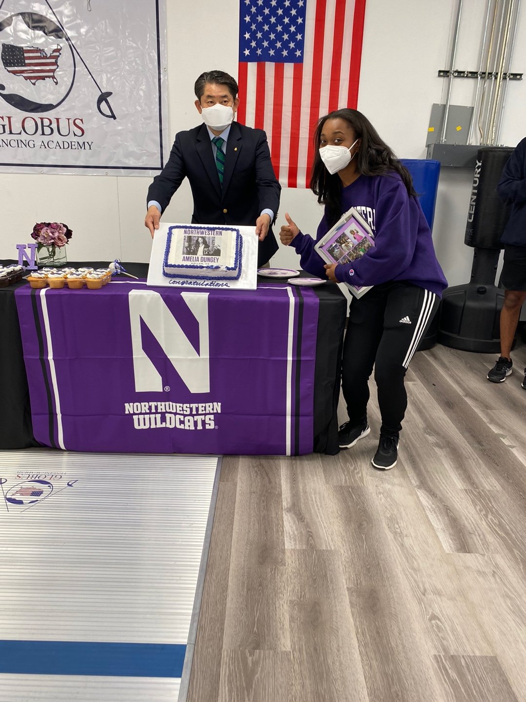 A young woman and a man, both wearing face masks, standing at a table with a purple Northwester Wildcats banner. The man is holding a cake with a photo and the words 'Northwestern Amelia Dungey', celebrating her achievement. The table has cupcakes an