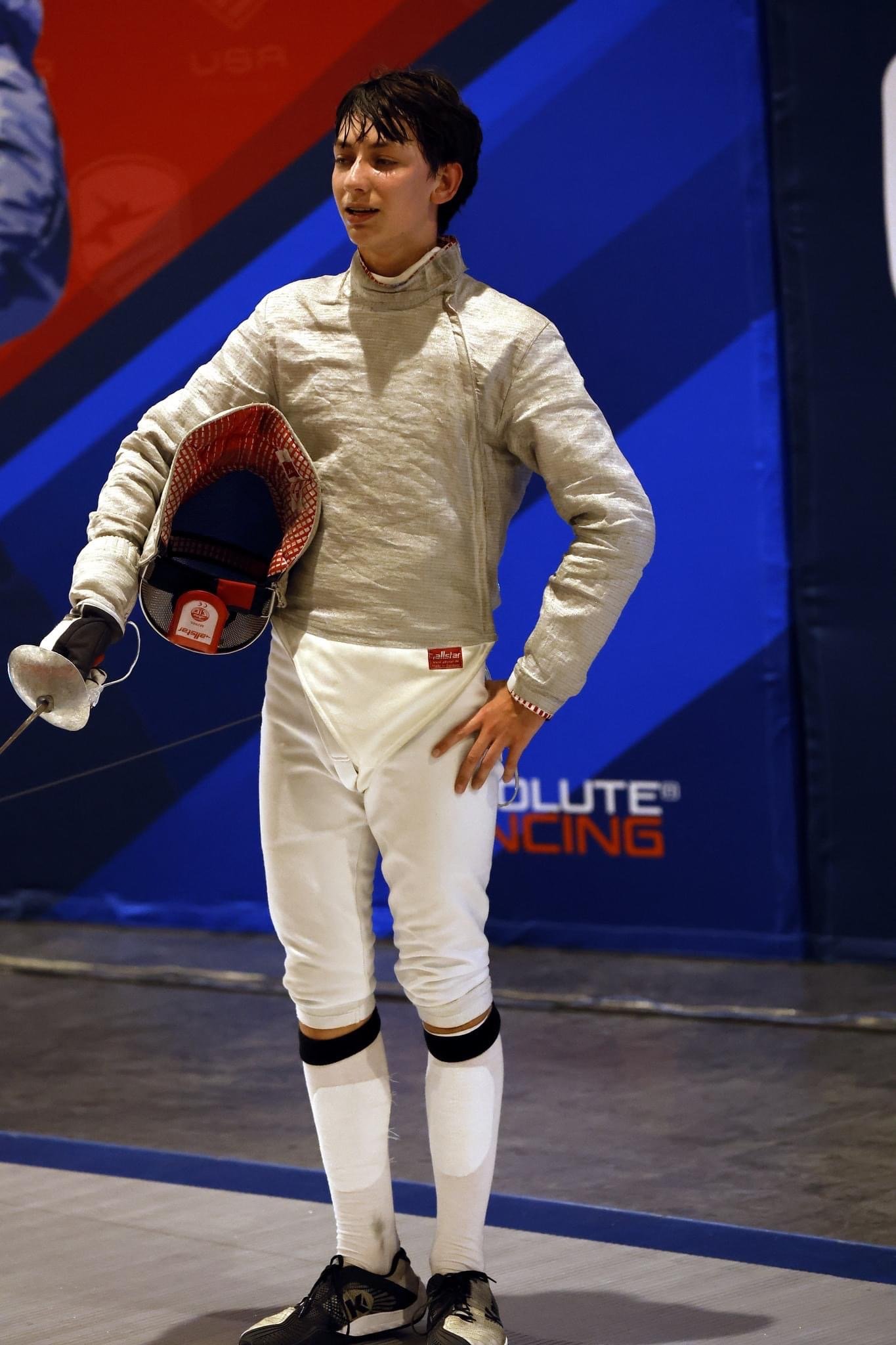 Young male fencer standing on a fencing strip, holding his fencing mask in one hand, wearing a fencing outfit and sports shoes, with a blue and red background that has the words "Absolute Fencing".
