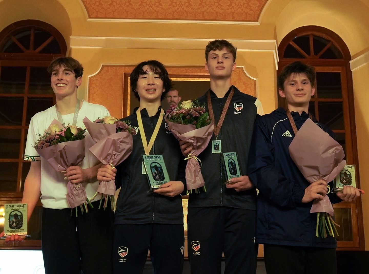 Four young men standing on a podium holding bouquets and awards, with medals around their necks, during an award ceremony in an elegant room with arched windows and a large mirror behind them.