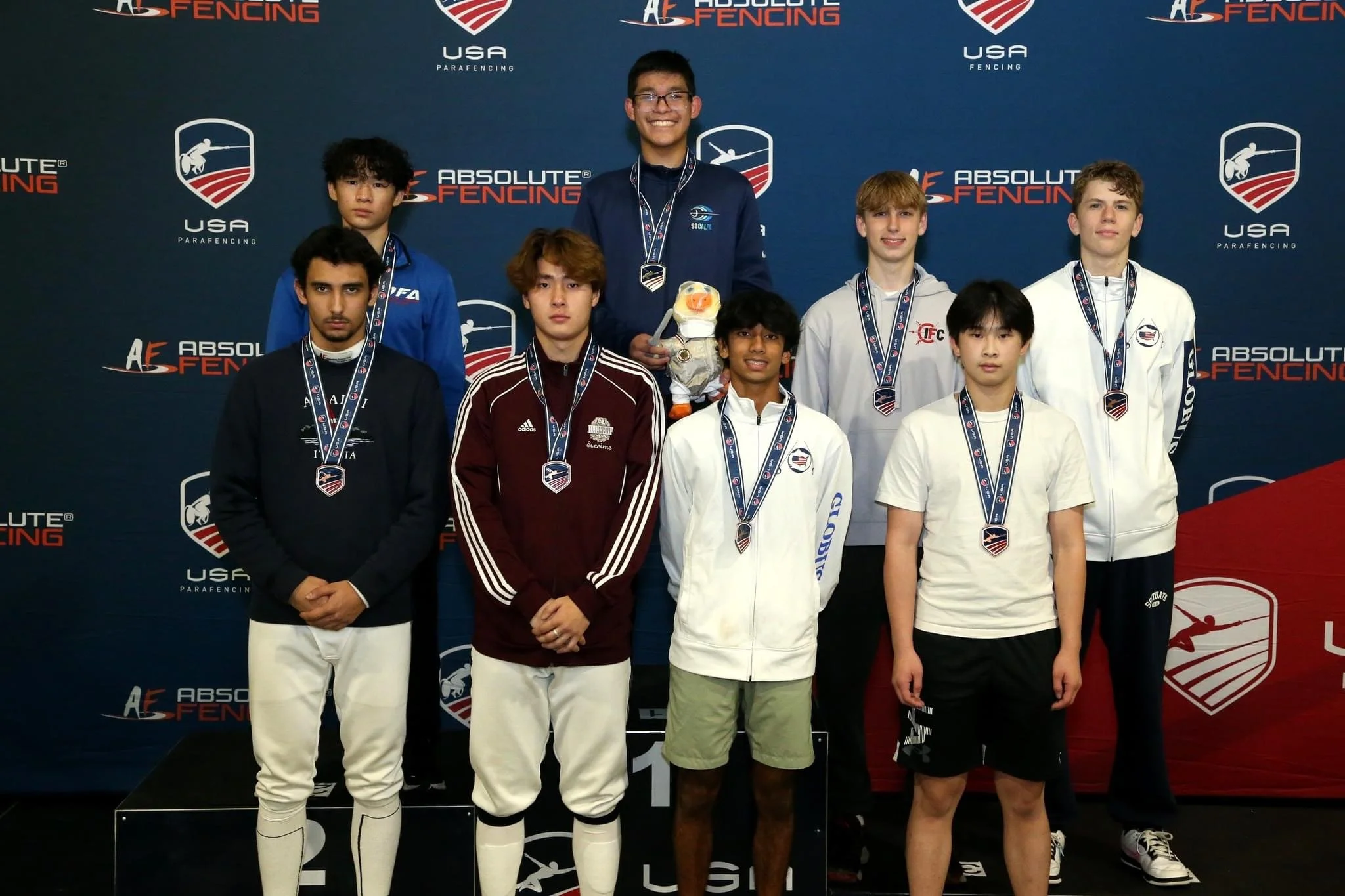 Young male fencers standing on a winners' podium with medals at a USA Paral fencing event, posing for a group photo in front of a blue backdrop with logos.