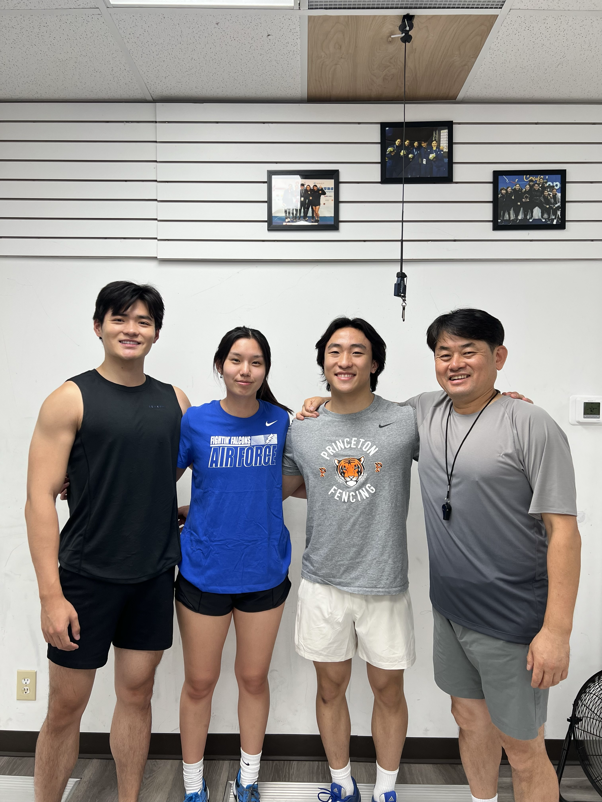 Four people standing together in a room, smiling, with a white wall behind them and framed photos hanging on the wall.