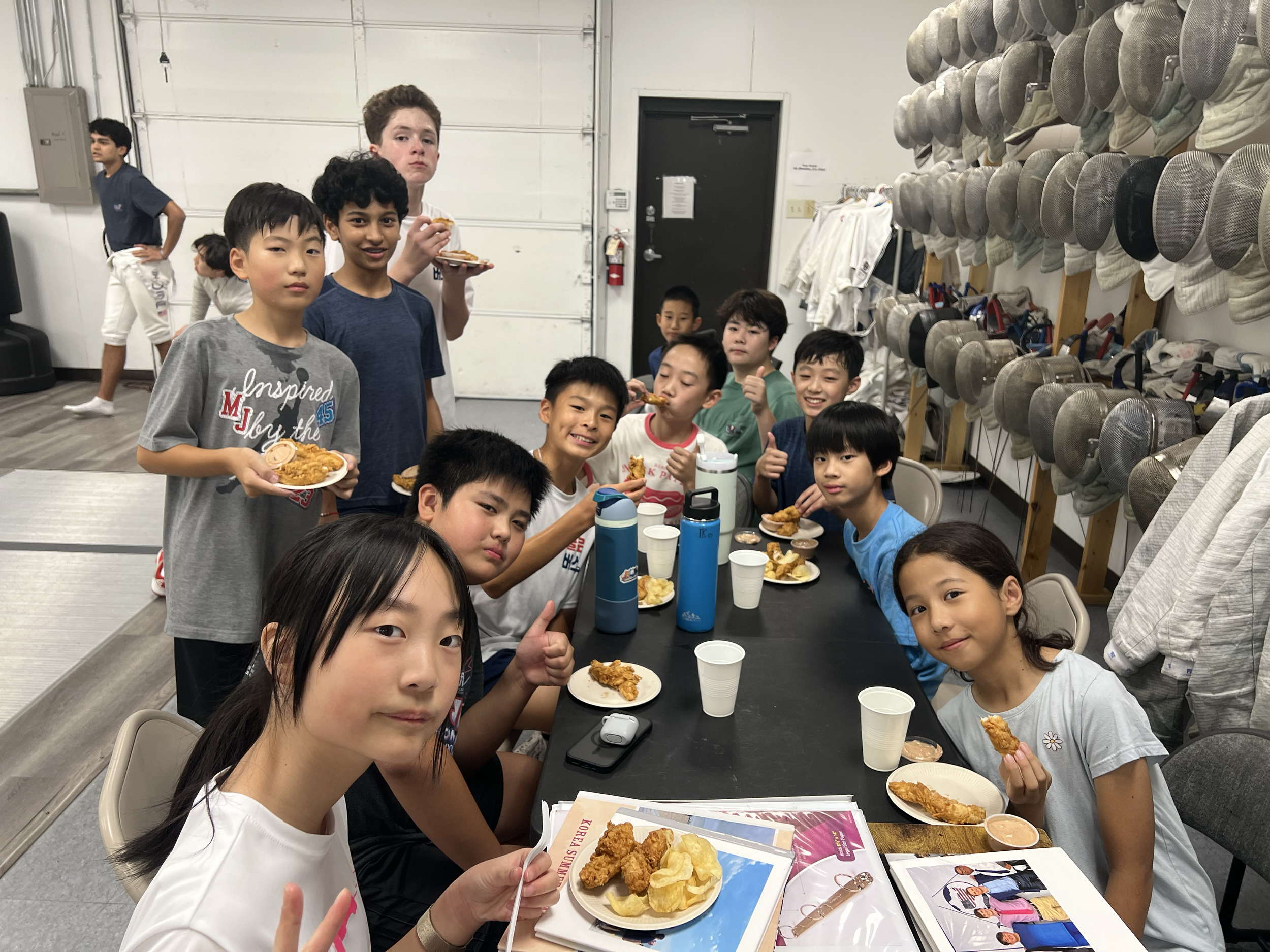 A group of children sitting and standing around a table, eating fried chicken, with some holding plates and giving thumbs up, in a room with fencing gear on the wall.