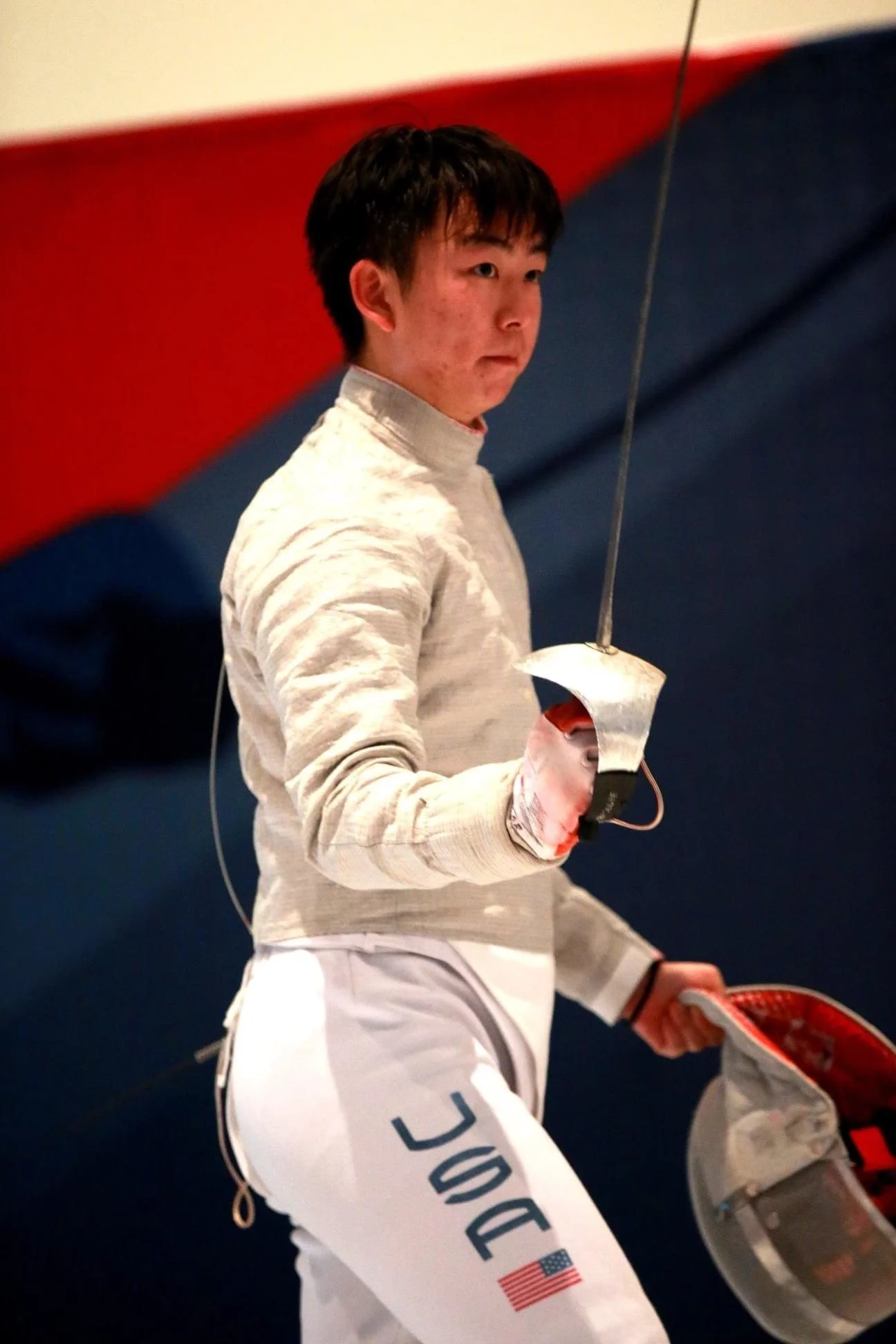 A male fencer in fencing gear holds a foil and wears a mask in his hand.