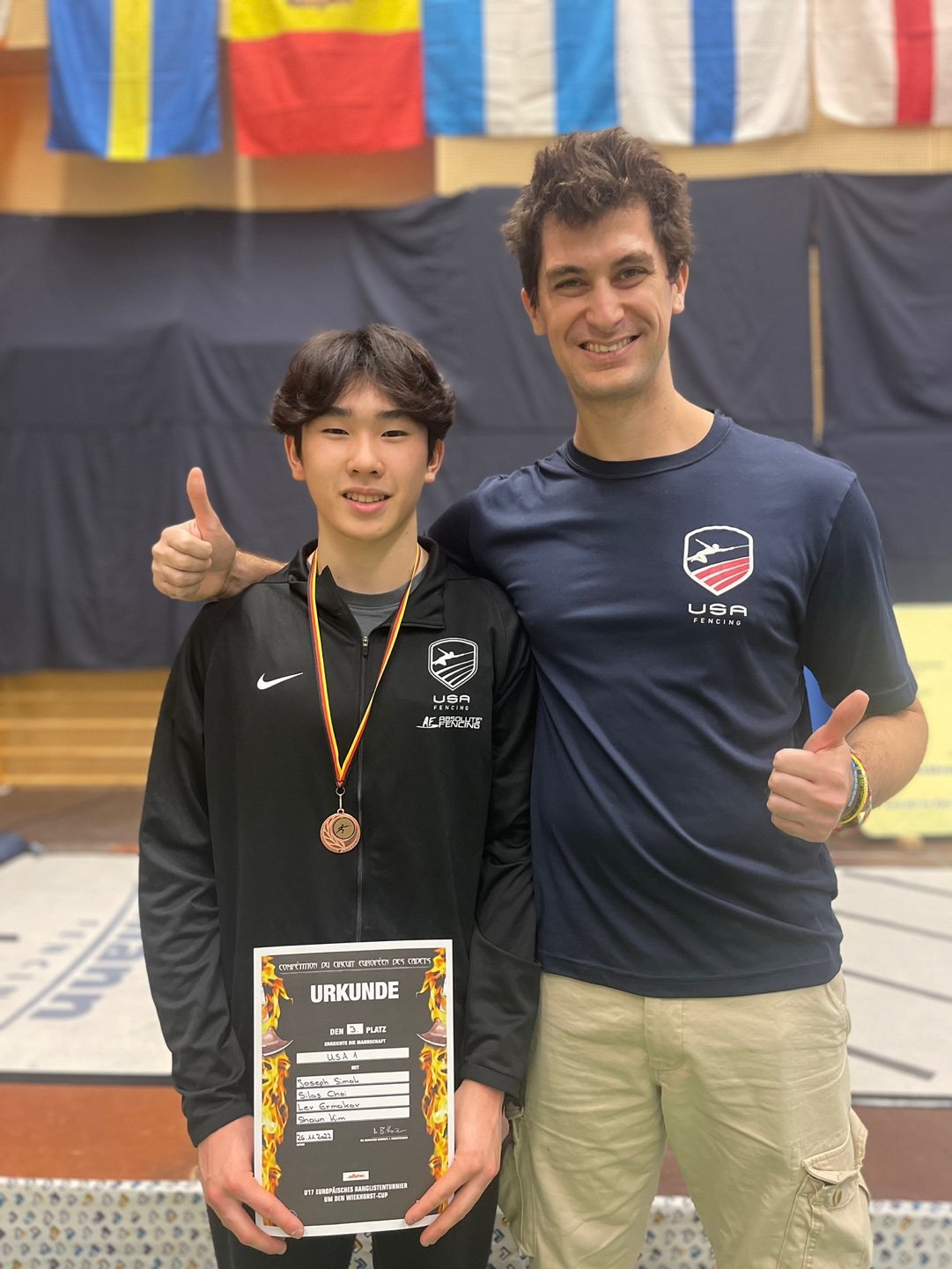 A young male athlete in a black USA fencing jacket holding a certificate and wearing a medal, standing next to an older man in a blue USA fencing t-shirt, both giving thumbs up in an indoor sports venue with flags hanging from the ceiling.