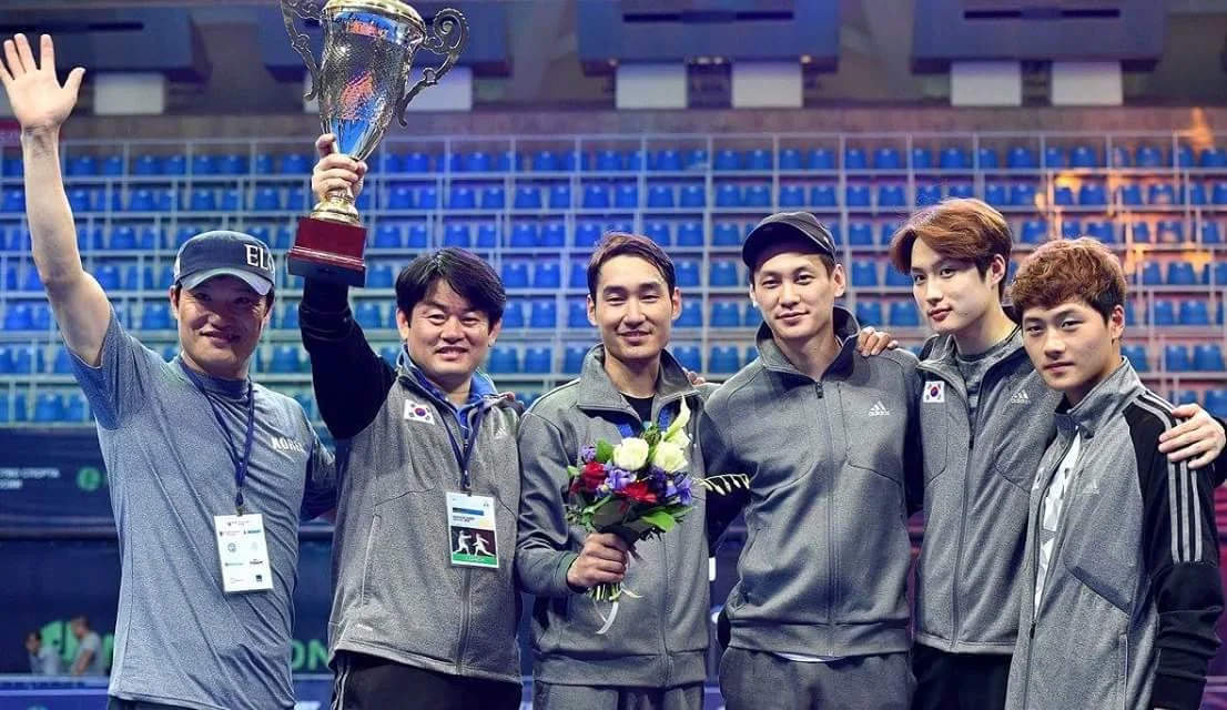 A group of six athletes standing together in an arena, celebrating with a trophy and a bouquet of flowers after winning a competition.