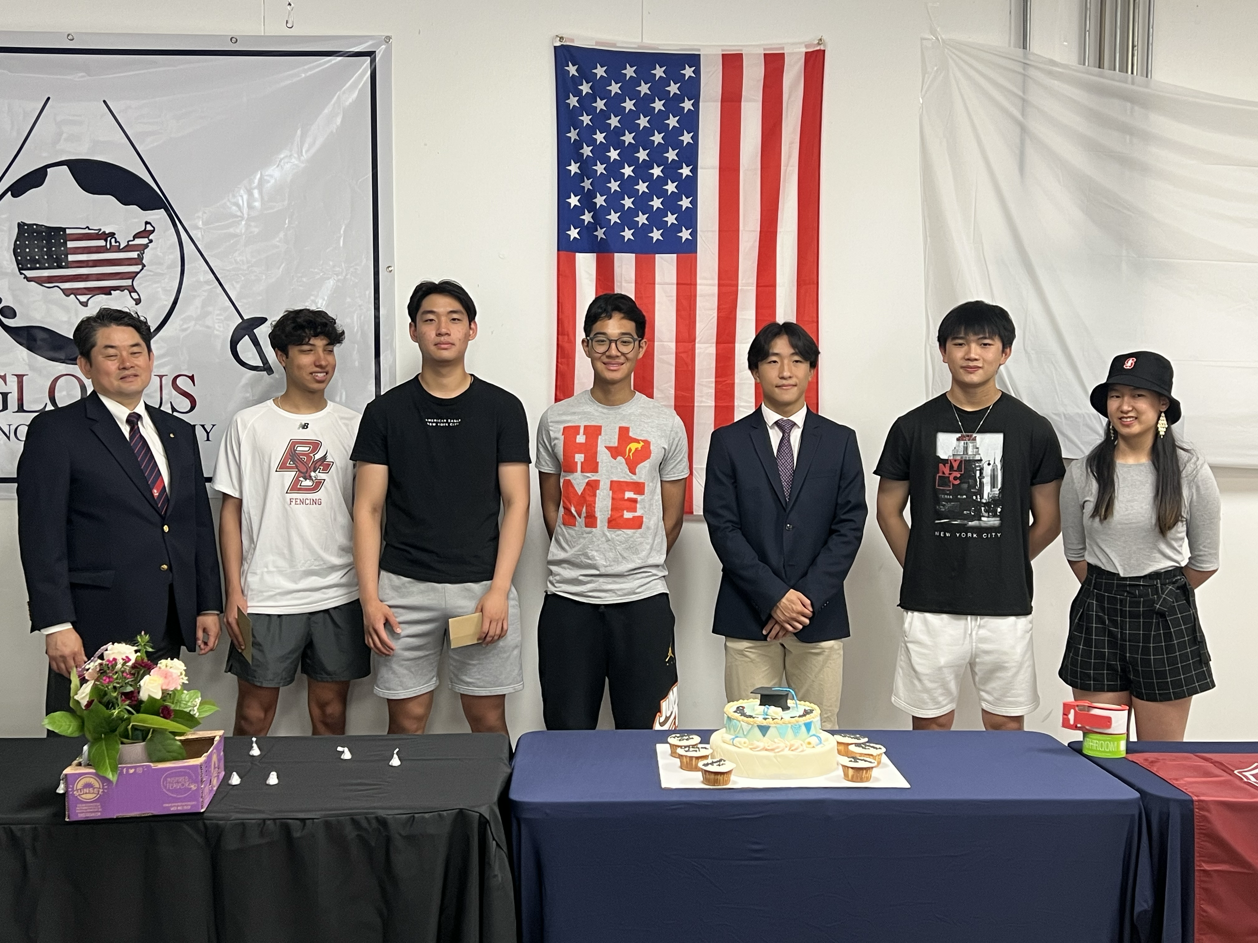 Group of seven young people and one adult, standing behind a table with a birthday cake and cupcakes, in front of American flags and a globe flag, at a celebration or ceremony.