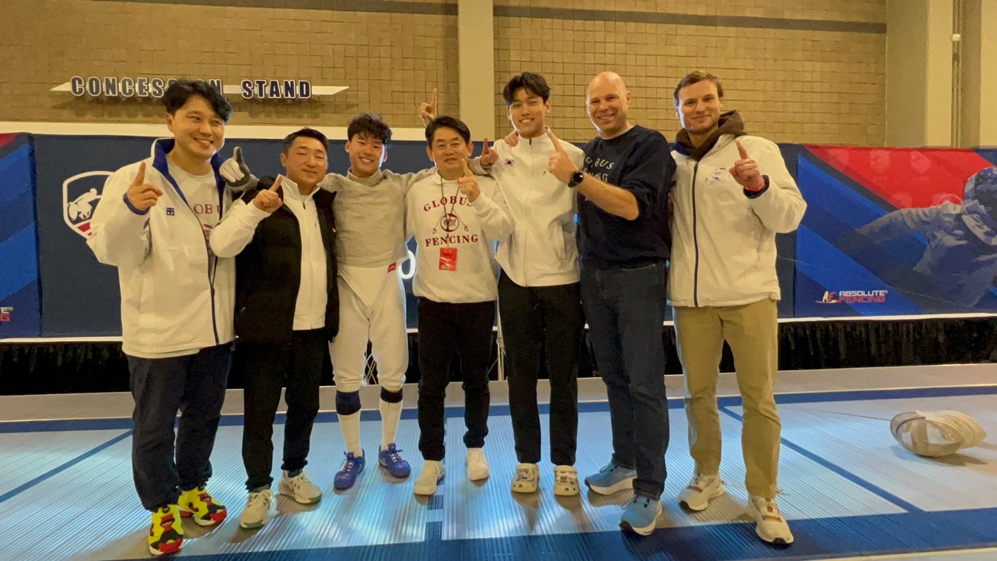 Group of seven fencers and their coach posing together at a fencing competition, all making thumbs-up gestures, in front of a backdrop with fencing-related graphics.