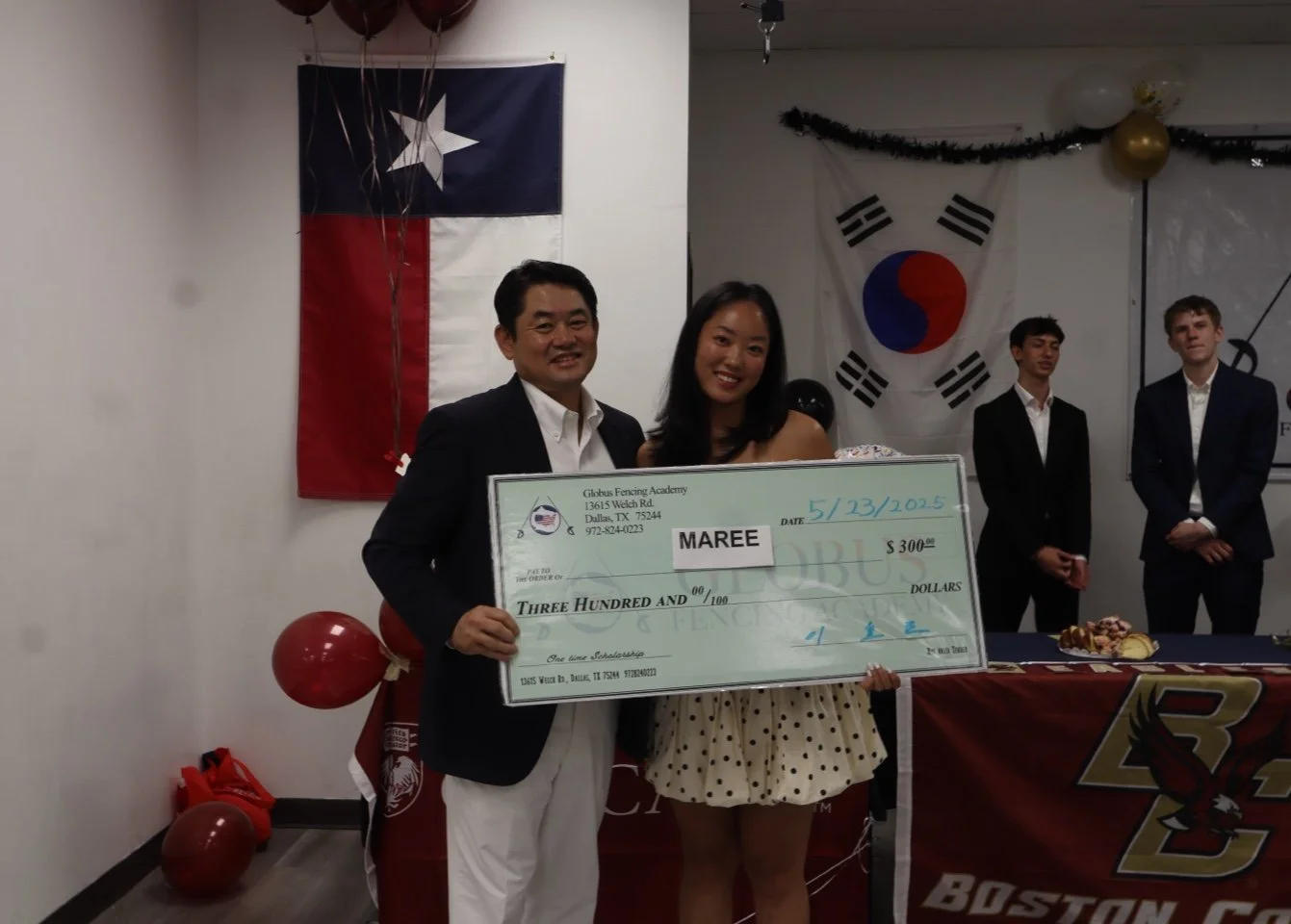 Two people, a man and a woman, holding a giant check during an award ceremony, with Texas and South Korea flags hanging in the background, and three other men standing nearby.