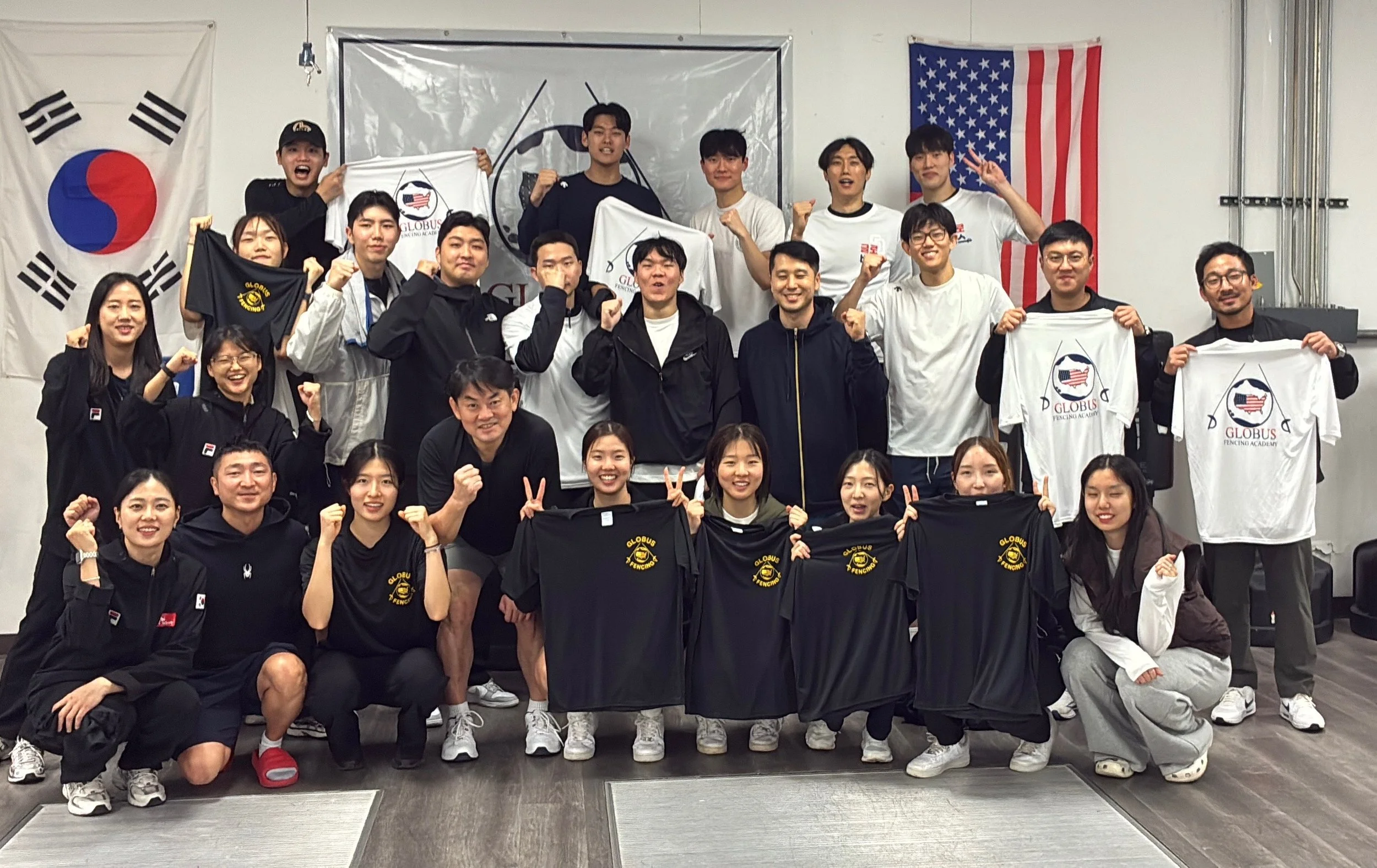 Group of young people posing with clenched fists, holding black and white T-shirts with gold lettering, in front of flags from South Korea and the United States, indoors.