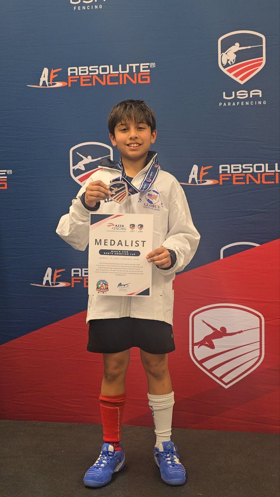 Young boy in fencing uniform holding a medal and a certificate, standing in front of a blue and red backdrop with fencing logos and text, smiling after a fencing competition.