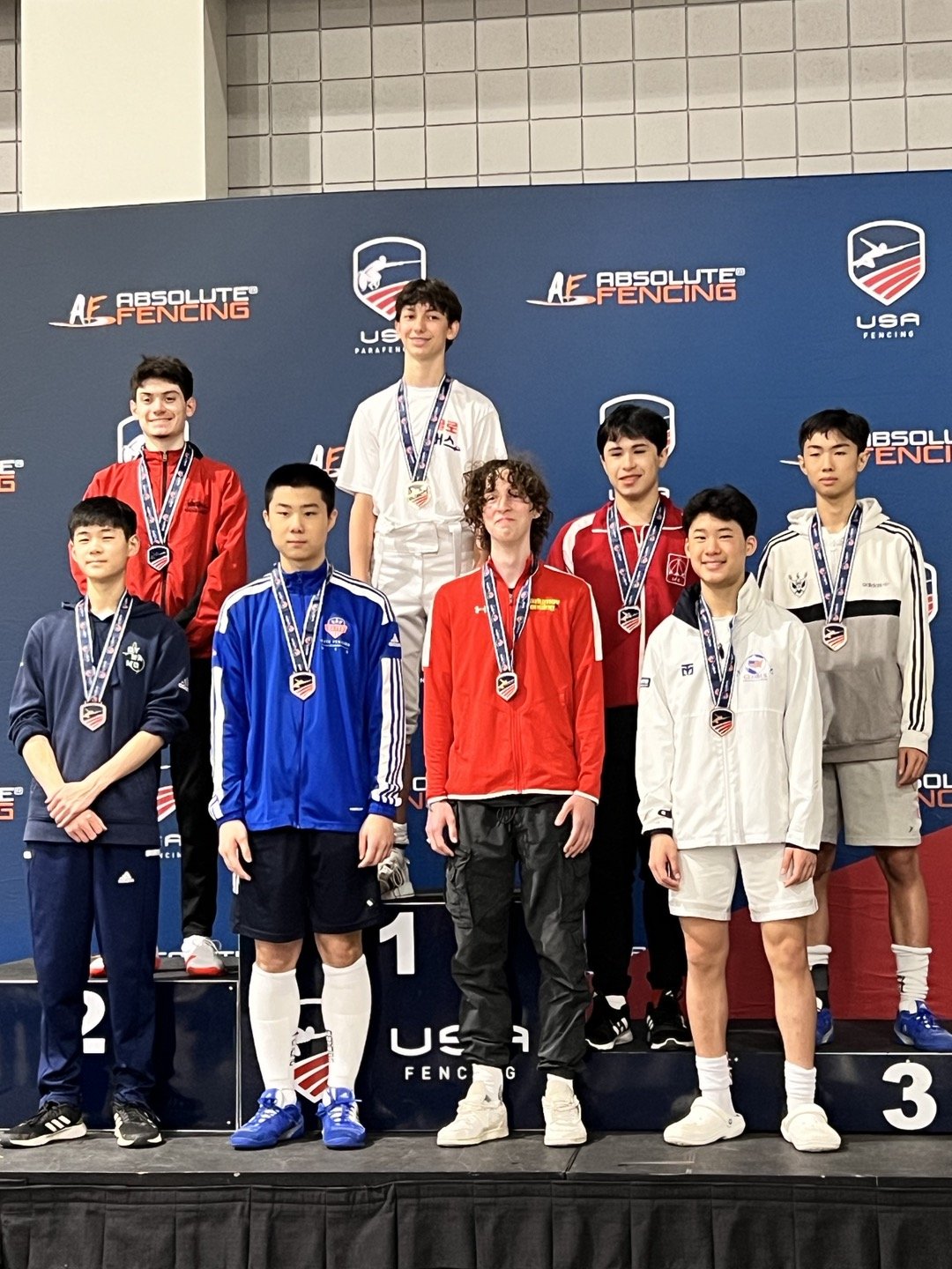 Young male fencers on the winners' podium at a USA Fencing event, wearing medals and sports jackets, with a backdrop displaying USA Fencing logos.