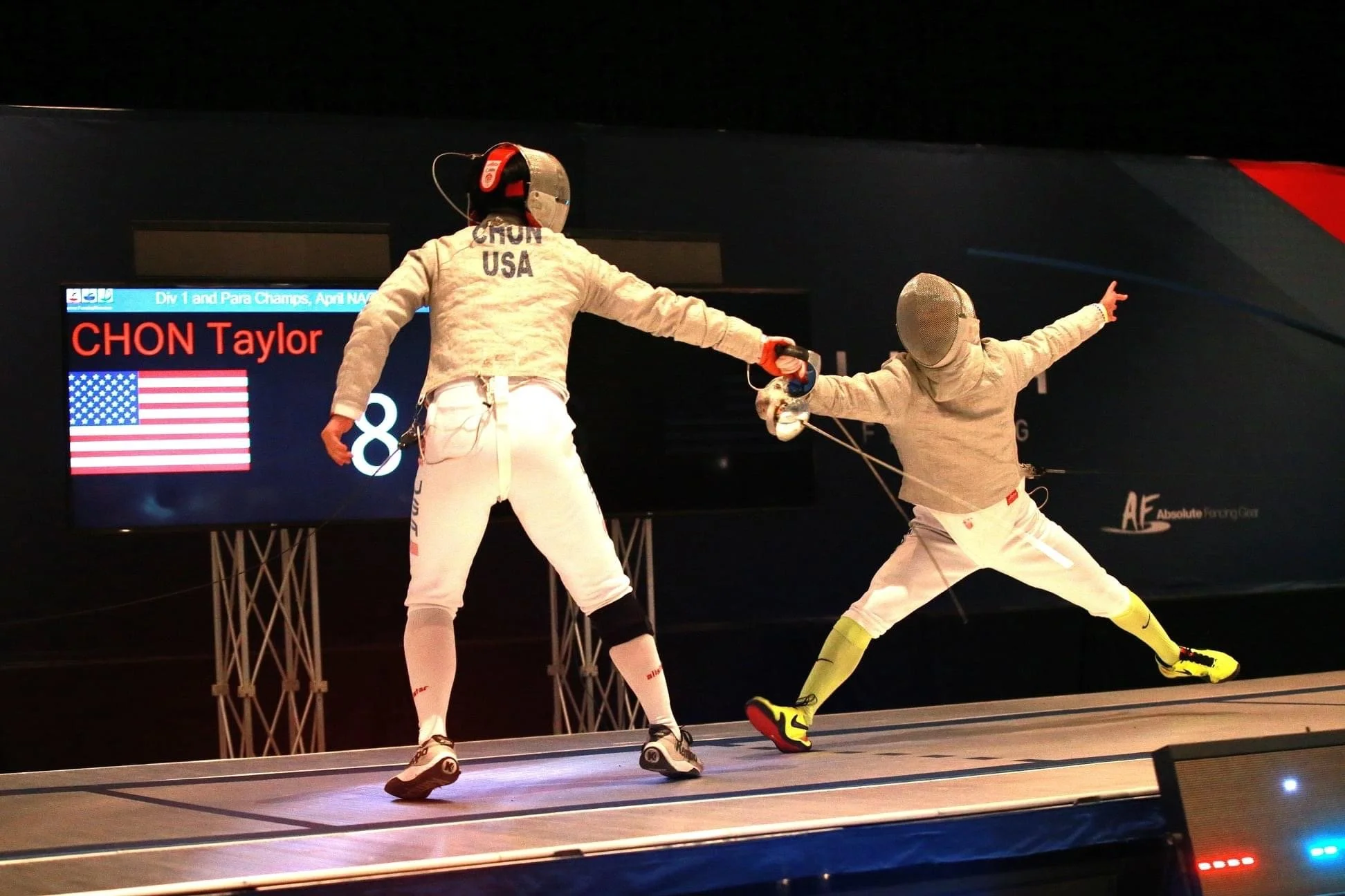 Two male fencers in protective gear engaged in a bout, with one prompting and the other lunging with a foil, on an indoor fencing strip during a competition.