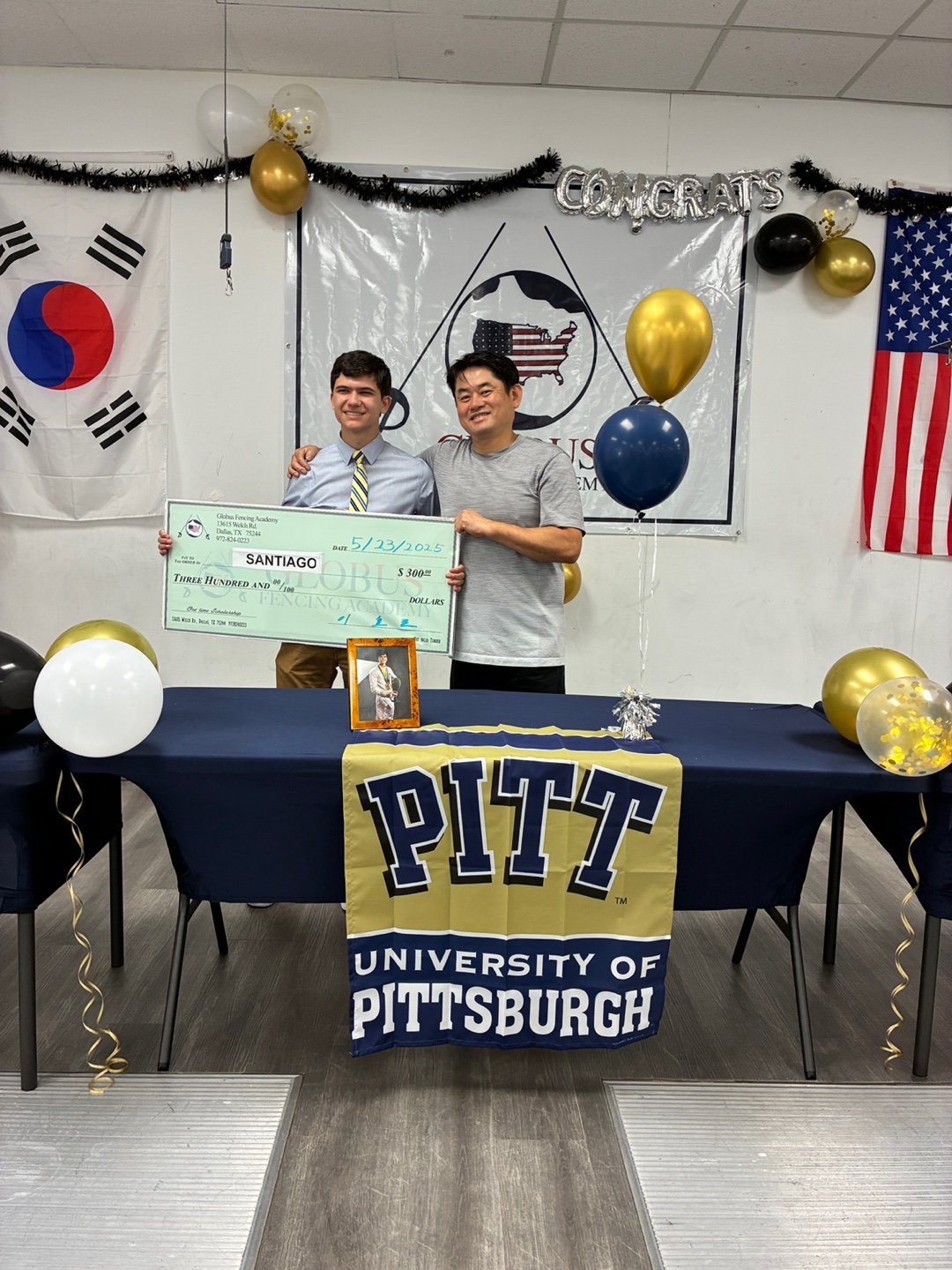 A young man and an older man standing behind a table with a Pitt University banner, holding a large check for $300, celebrating a fencing achievement, with balloons and congratulatory decorations in the background.