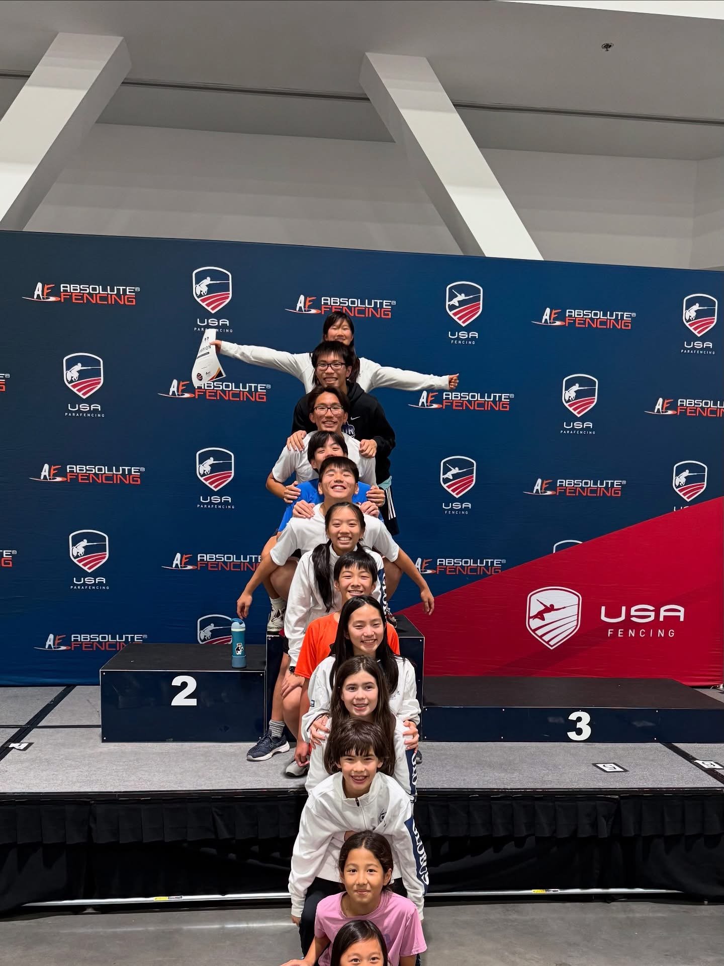 A group of young athletes standing in a line on a winners' podium, with some standing on the second and third place spots, at an indoor fencing competition. All are smiling, and there is a blue backdrop with logos of USA Fencing and Absolute Fencing 